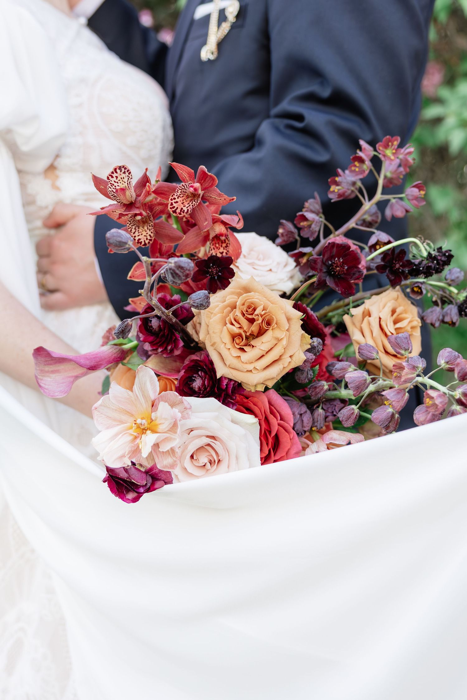 Stunning bridal bouquet featuring deep red, burgundy, peach roses, orchids, and dahlias held by a bride and groom.