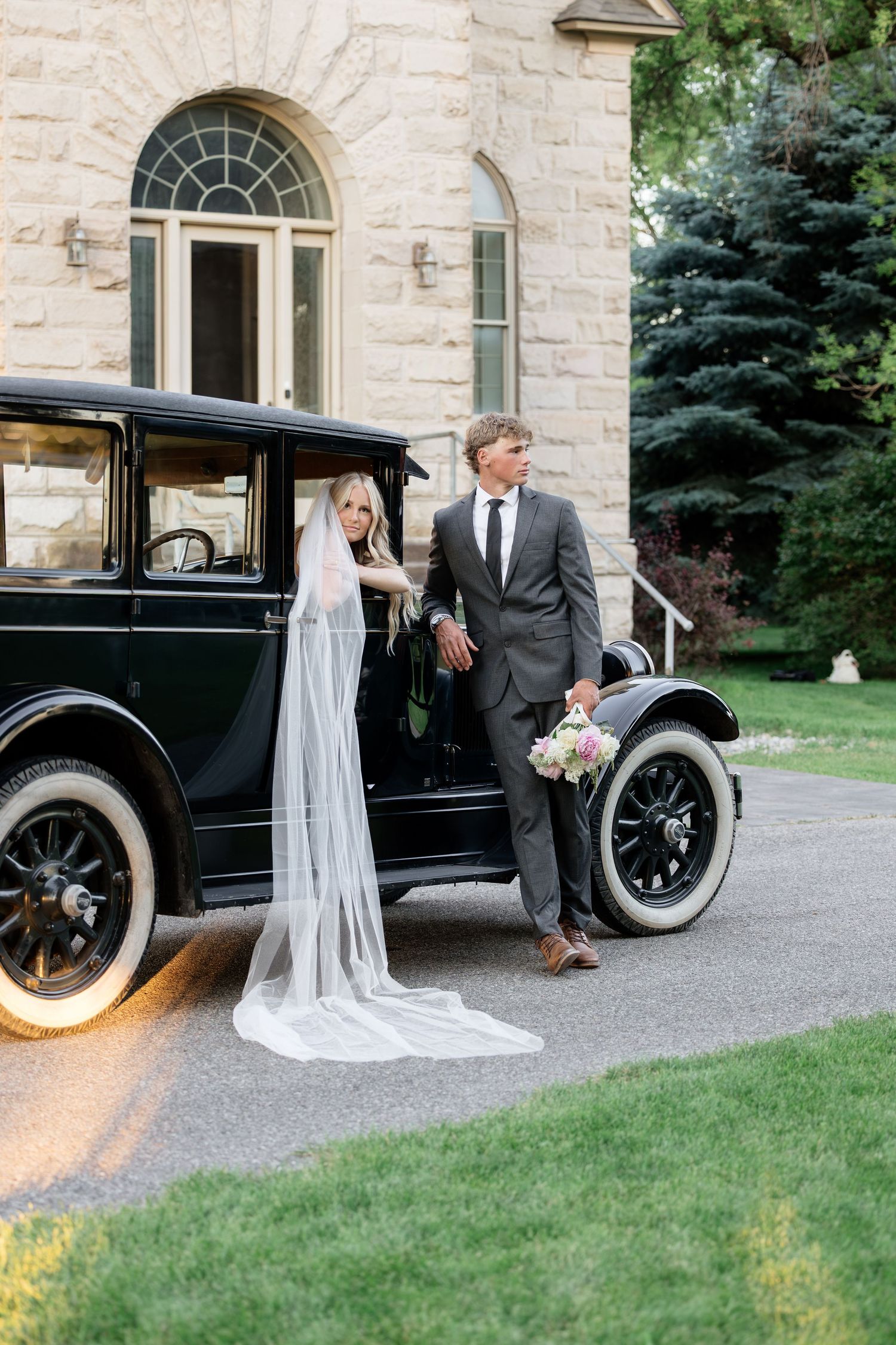 Bride and groom pose beside a vintage black car outside a stone mansion, bride wearing a flowing cathedral veil.