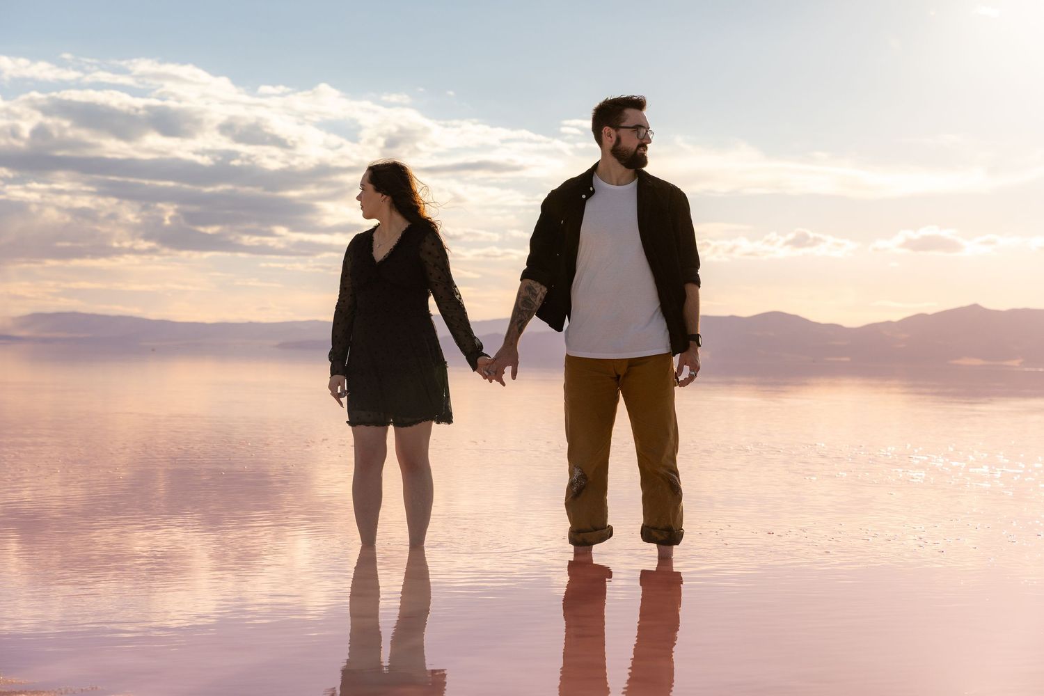 Couple holding hands standing on reflective water at sunset with mountains in the background.