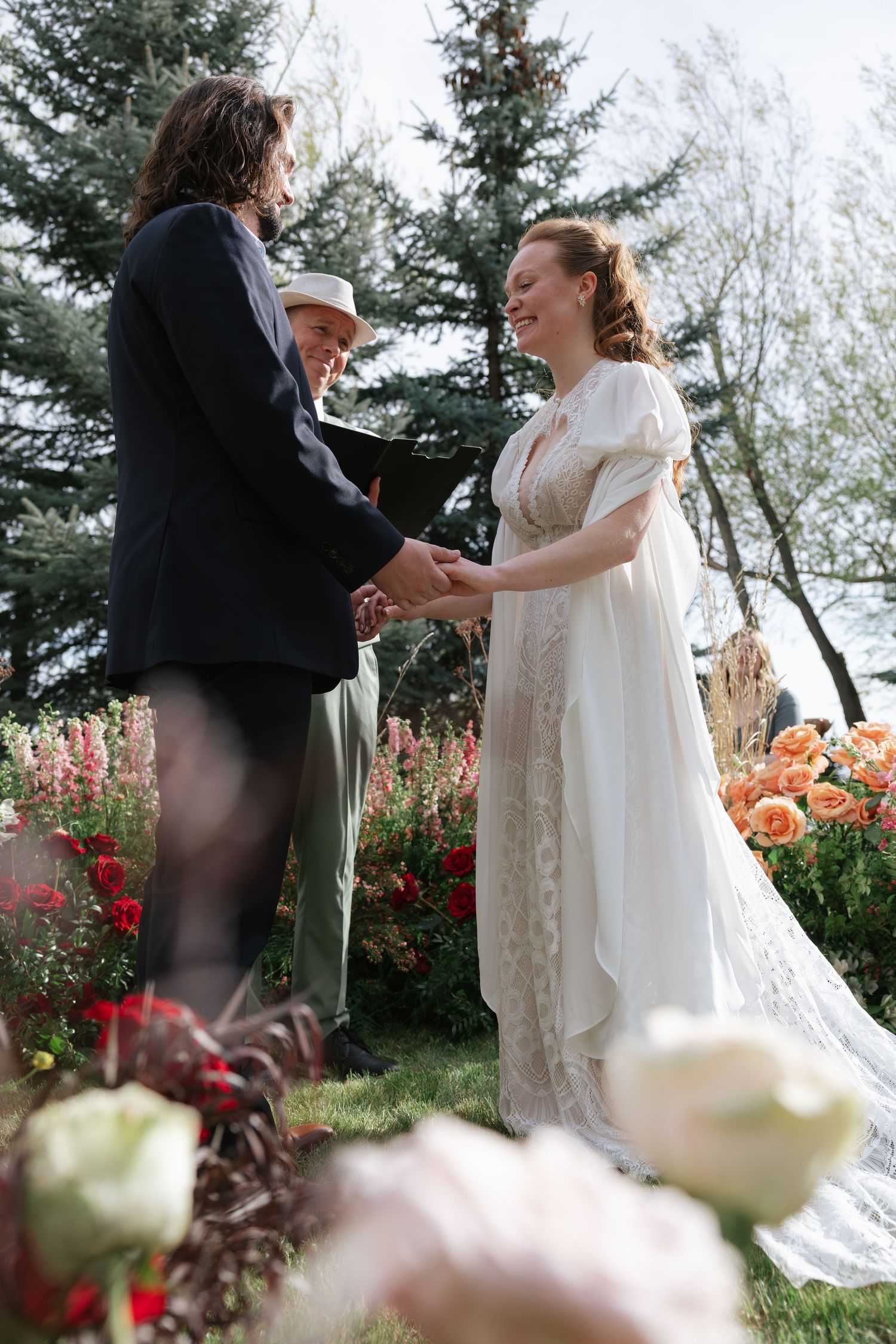 Bride in white lace gown holds hands with groom during outdoor garden wedding ceremony with colorful flowers.