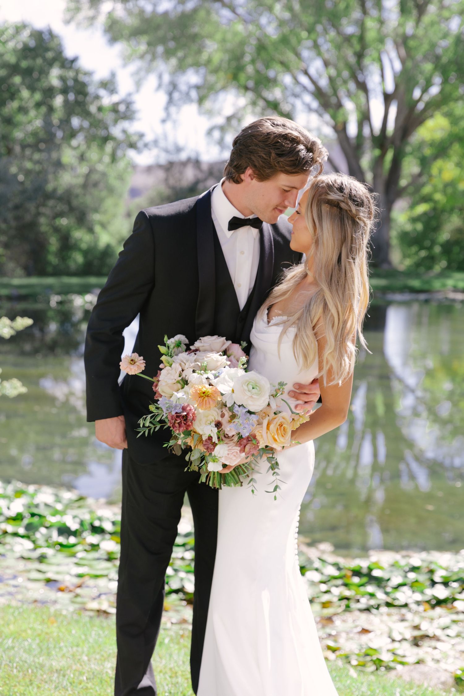 Bride and groom share a romantic moment by a lily pond, she holds a pastel bouquet in her white gown and he wears a black tuxedo.