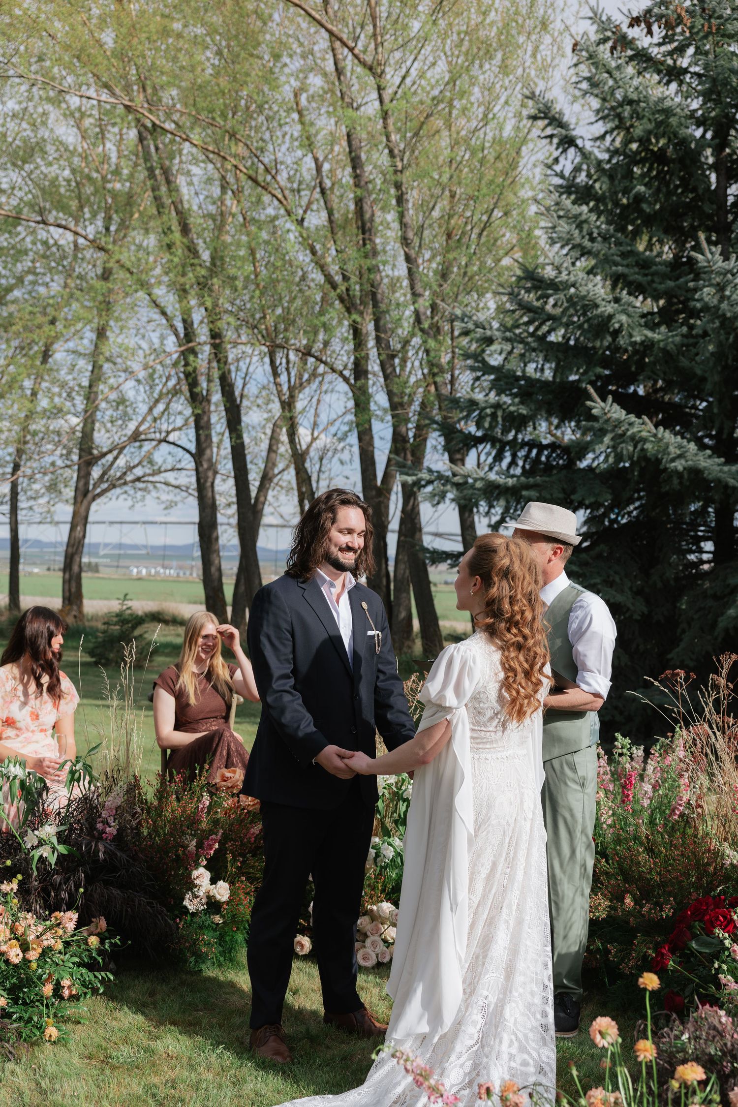 Couple exchanges vows at outdoor garden wedding ceremony surrounded by colorful floral arrangements and lush trees.