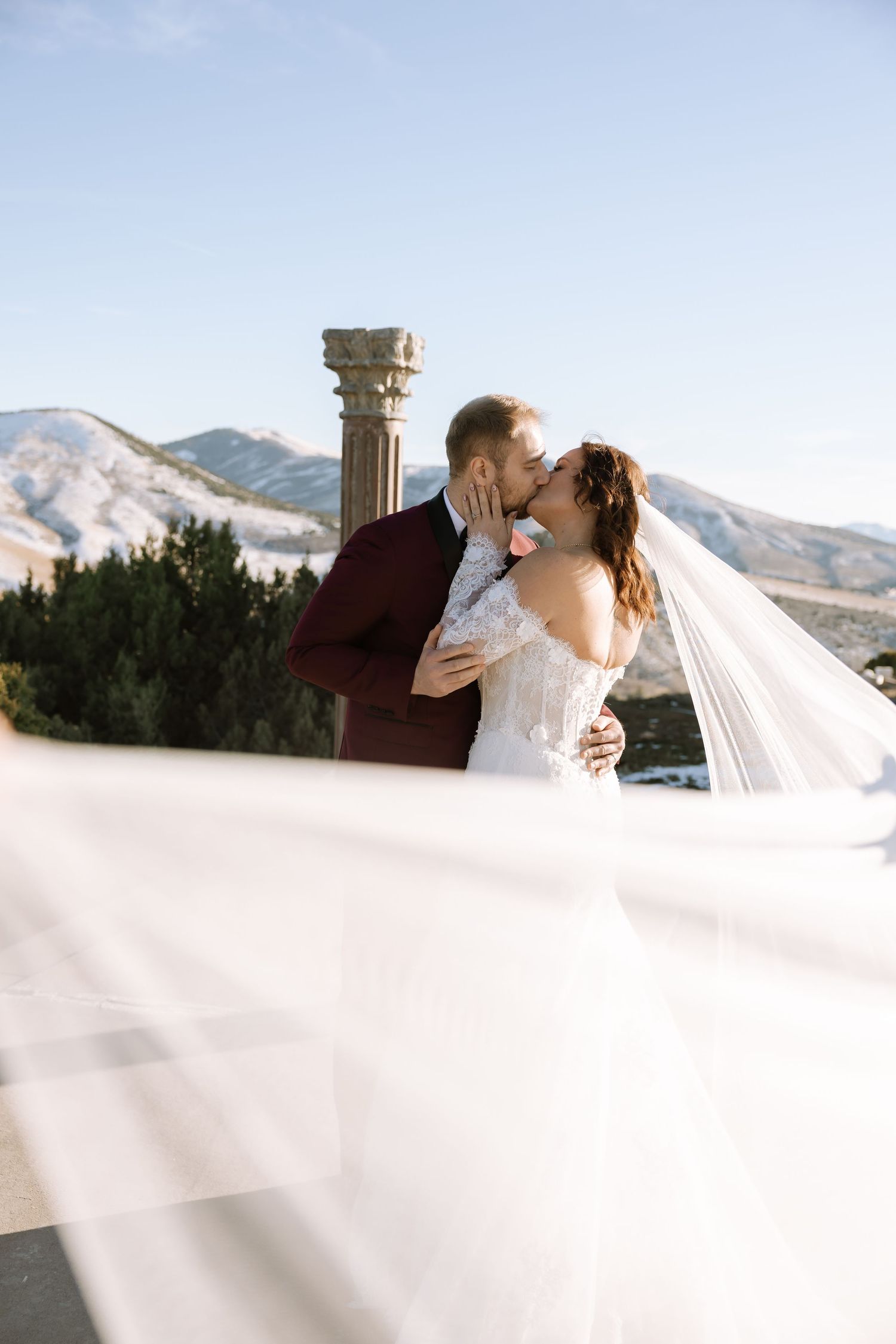 A romantic wedding moment captured against a mountain backdrop with a flowing white veil and stone pillar.