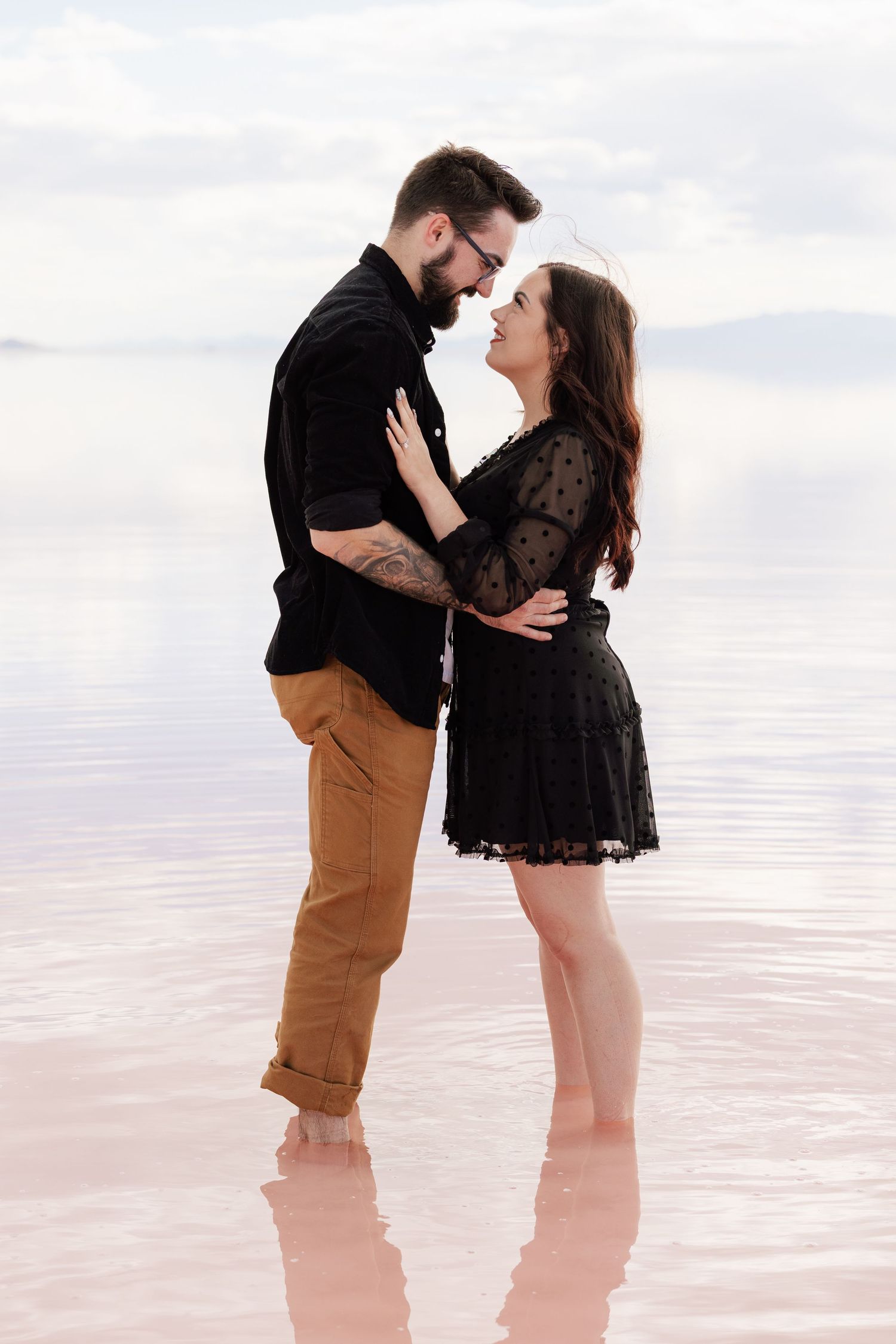 Couple in black outfits standing barefoot in shallow pink water, sharing a romantic moment with mountains in the background.