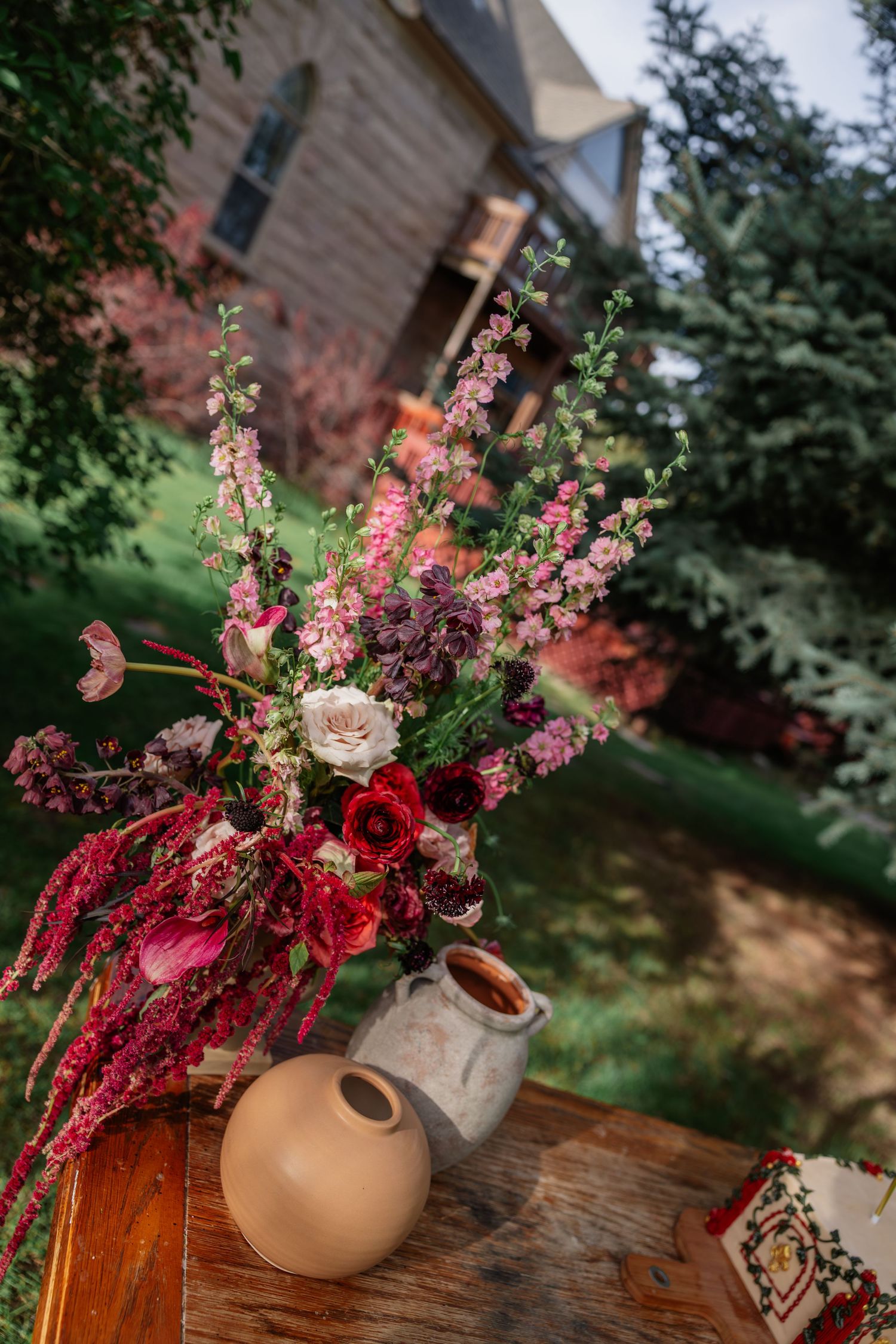 Rustic floral arrangement of red, pink and white blooms in ceramic vase on wooden table with house backdrop.