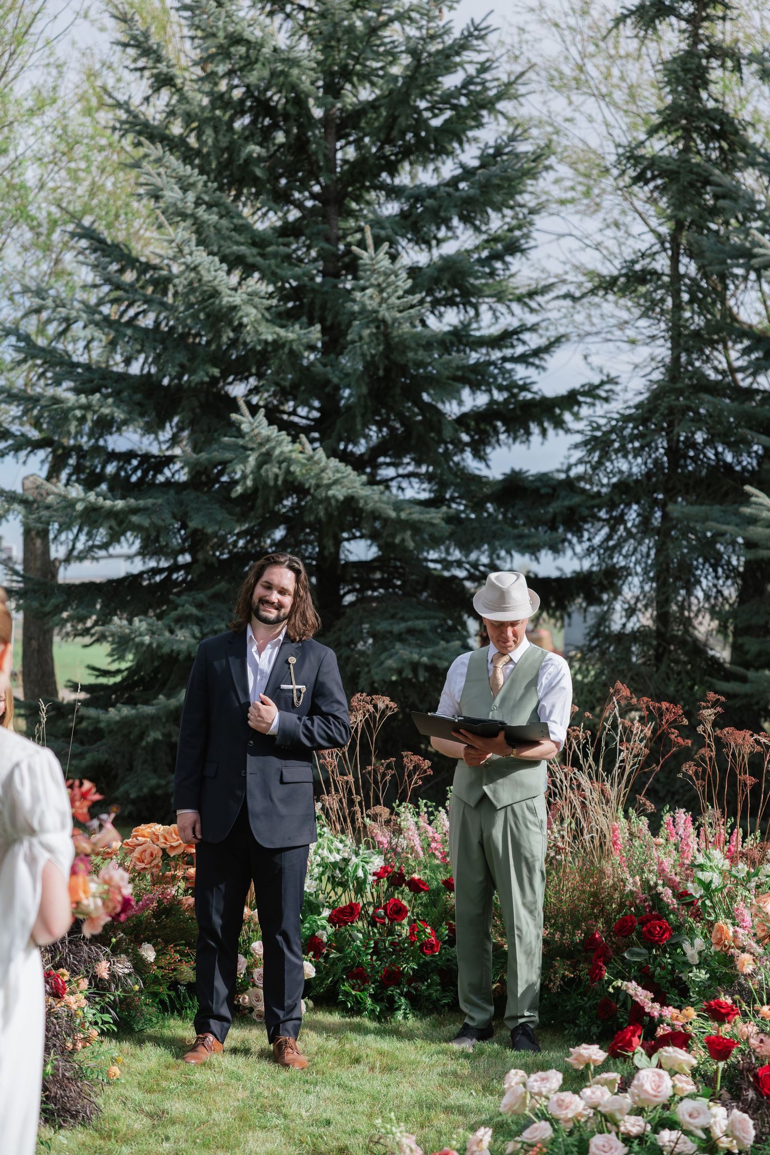 Groom in navy suit smiles at altar with officiant during outdoor garden wedding ceremony.
