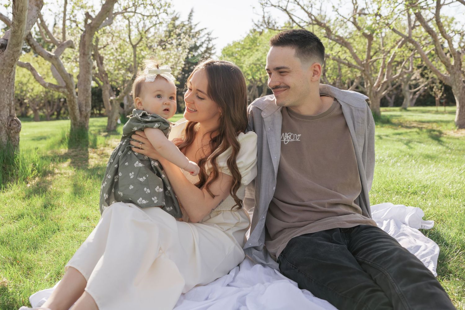 A young family sits on a blanket in a green orchard, with a mother and father smiling at their baby.