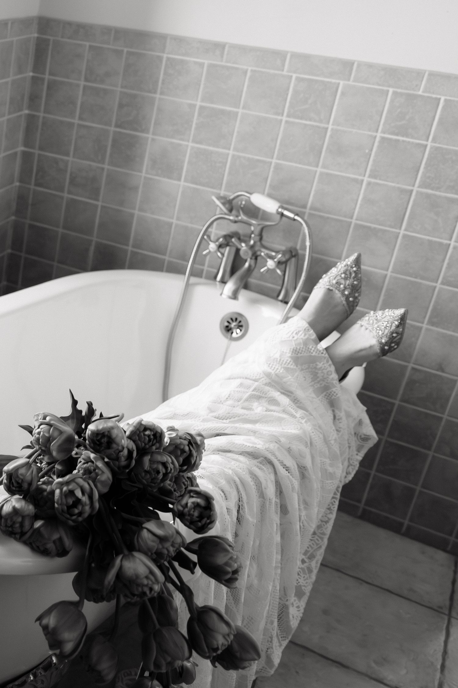 Black and white photo of bride resting in vintage clawfoot bathtub with bouquet and heels.
