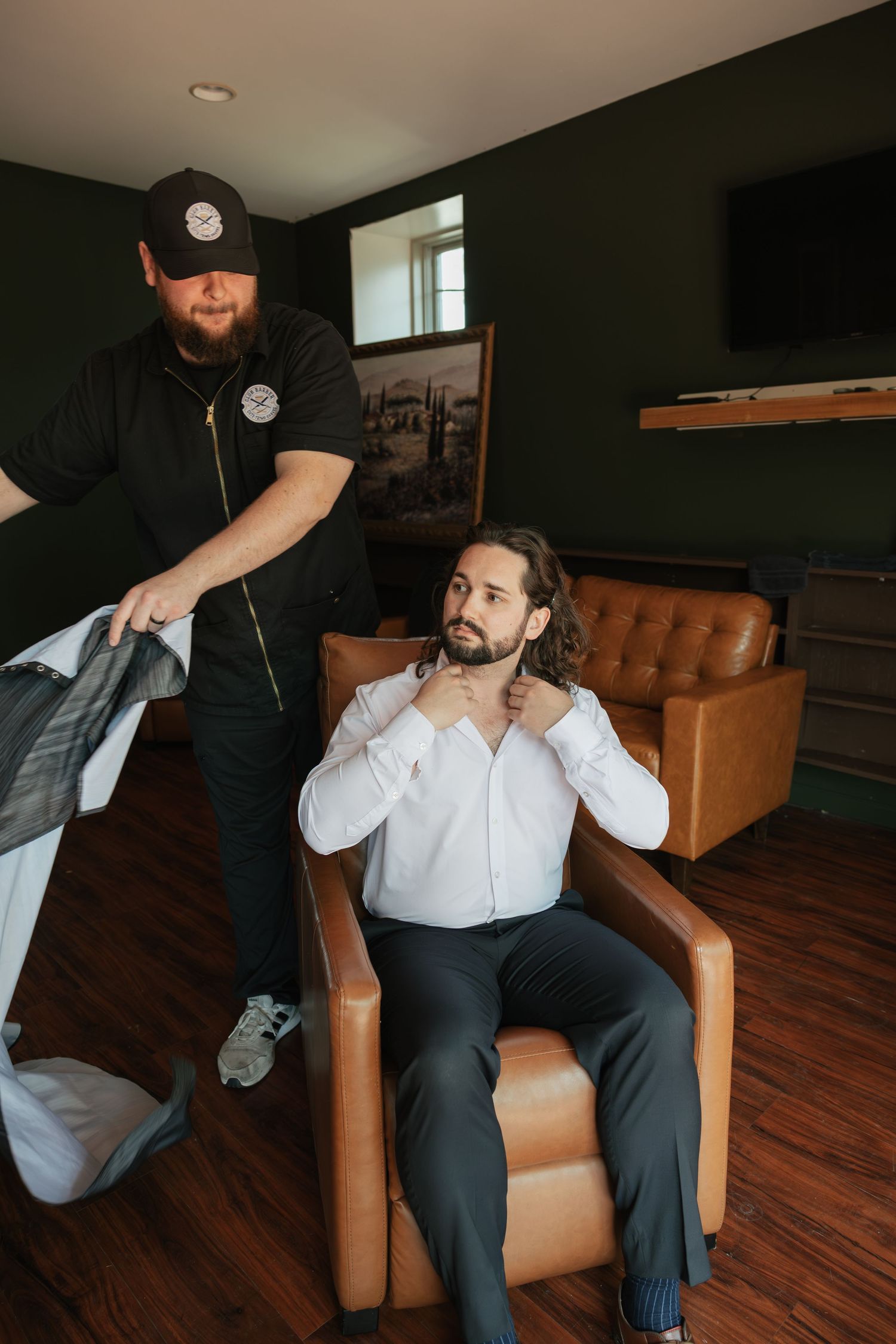 Groom in white shirt adjusting collar in leather chair while groomsman holds suit jacket indoors.