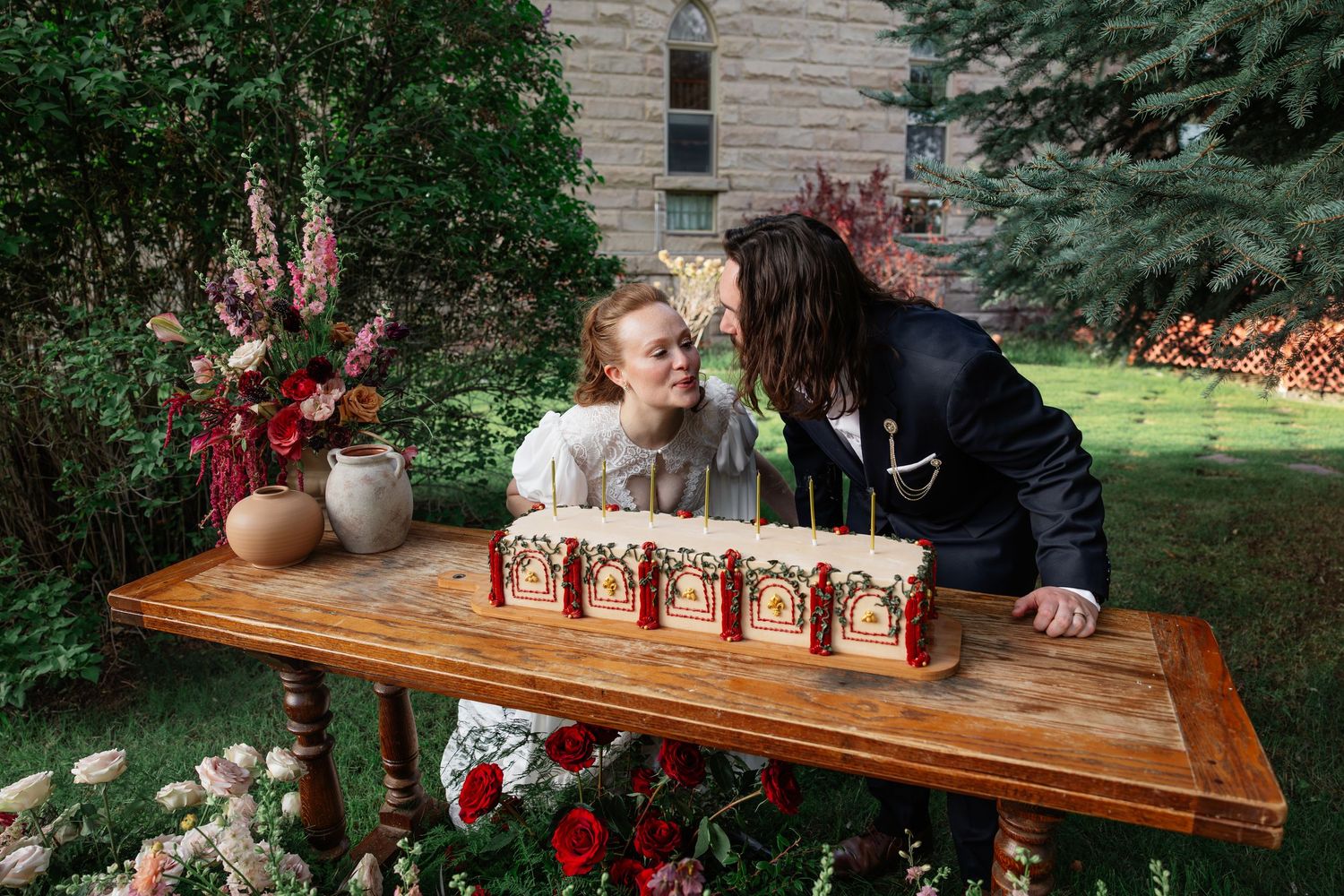 Bride and groom sharing a playful moment leaning in for a kiss over a decorative wedding cake on a wooden table.