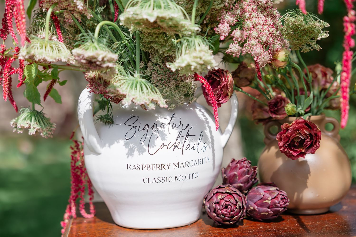 White ceramic pitcher displaying signature cocktails menu with lush floral arrangement of burgundy and green blooms.