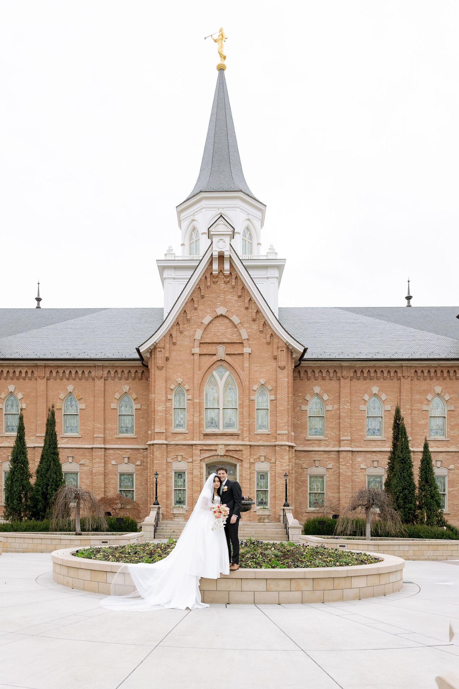 Bride and groom pose before a red brick LDS temple with white steeple and golden angel spire on their wedding day.
