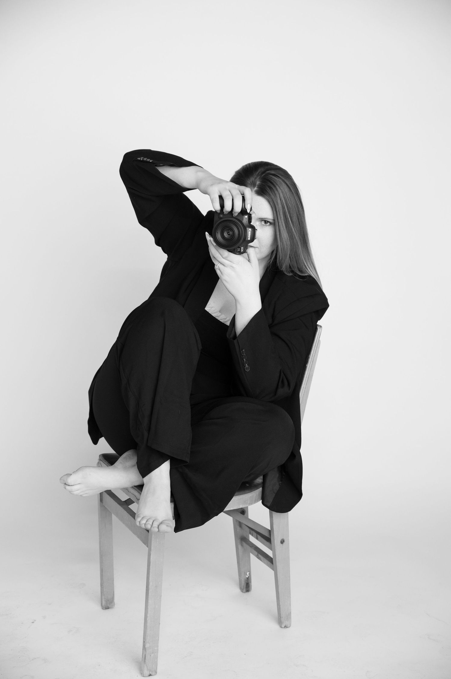 Black and white photo of a woman in black outfit sitting on a chair, holding a camera up to her face.