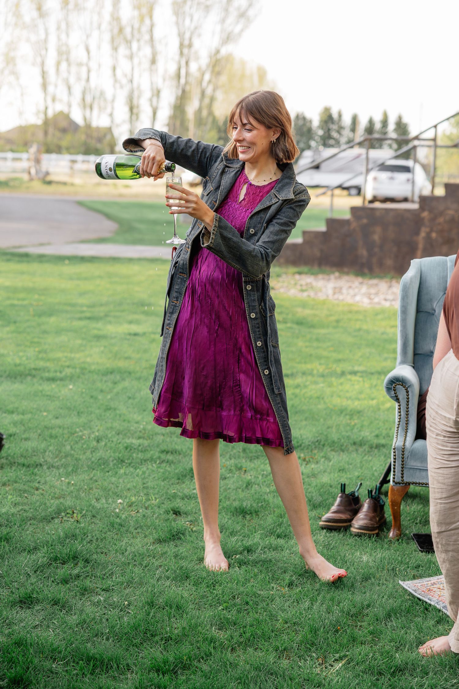 Smiling woman in purple dress and leather jacket pouring champagne barefoot on green lawn at wedding.