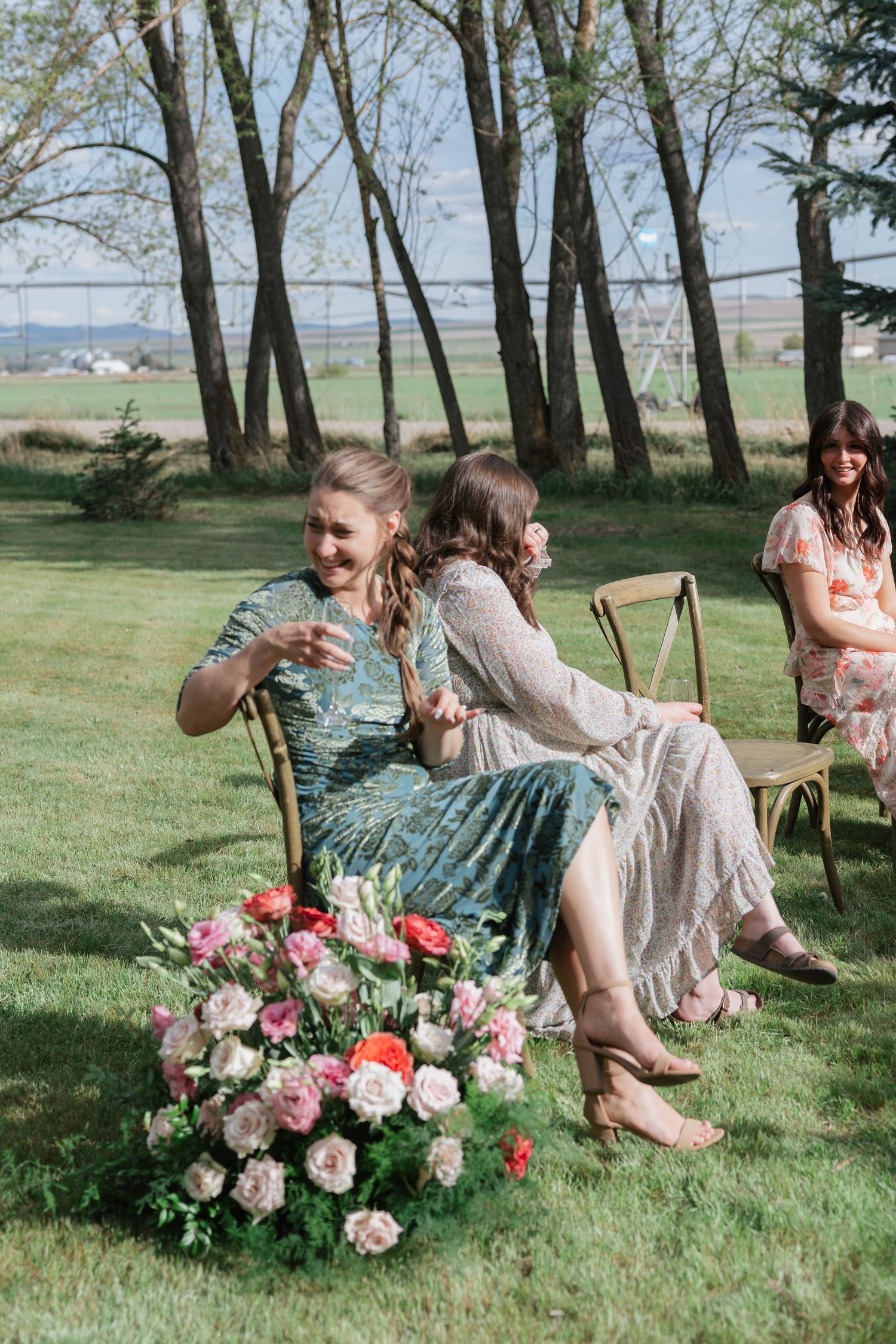 Two women in floral dresses sitting on wooden chairs at an outdoor wedding, laughing with pink roses at their feet.