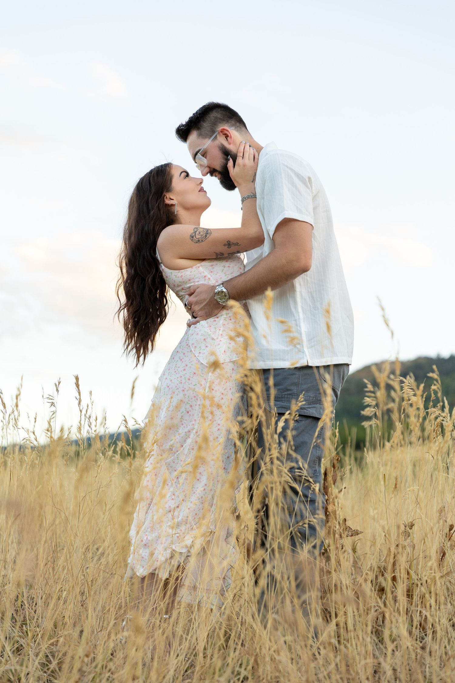 Romantic couple embracing in golden field, woman in floral dress, man in white shirt, about to kiss at sunset.