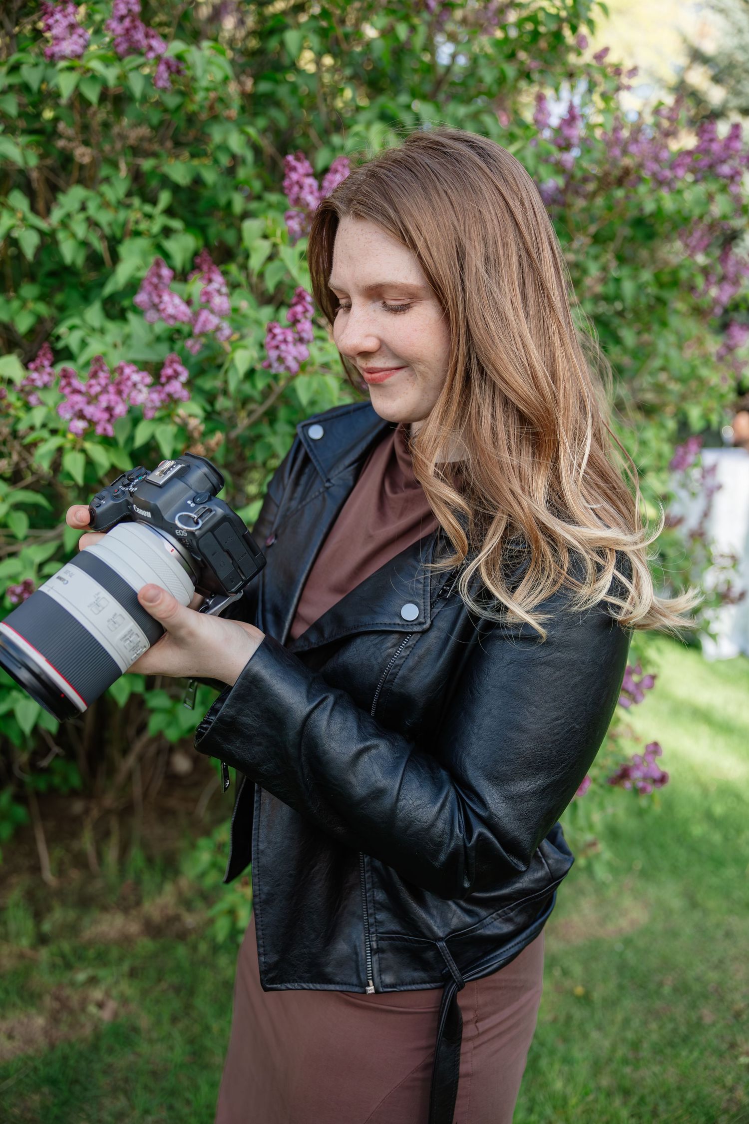 Young woman in black leather jacket smiling while reviewing photos on a DSLR camera outdoors near lilac bushes.