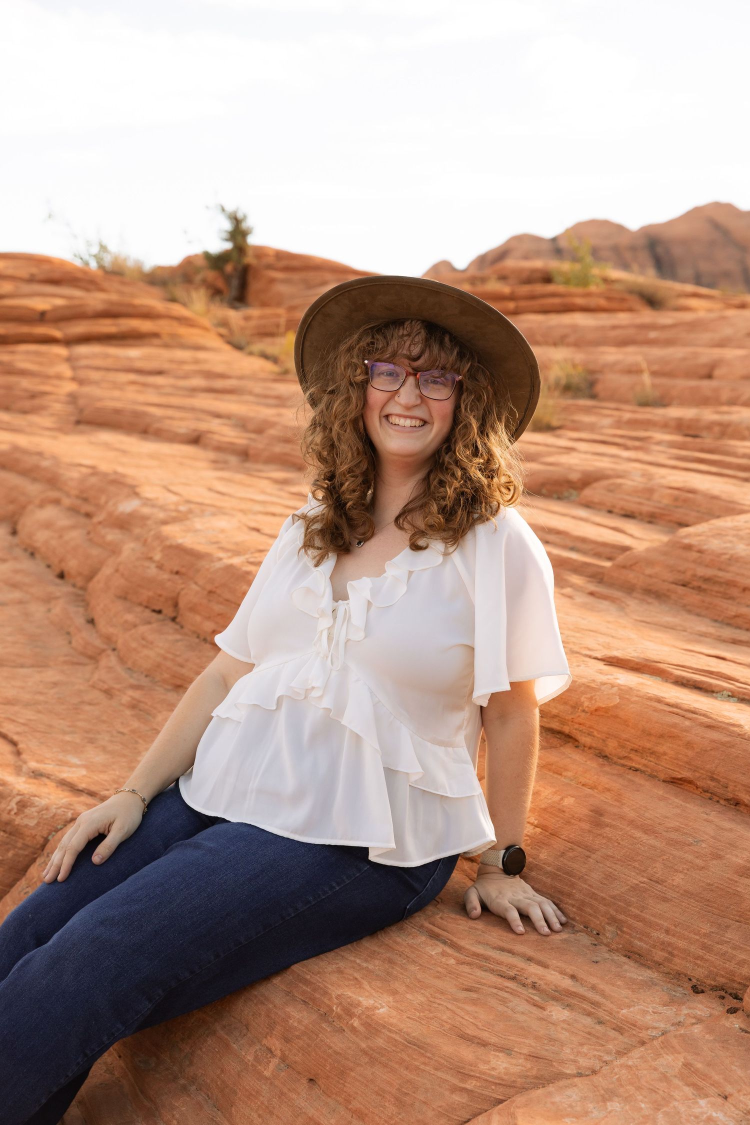 Young woman with curly hair, brown hat, and glasses sits smiling on red rock formations in a desert landscape.