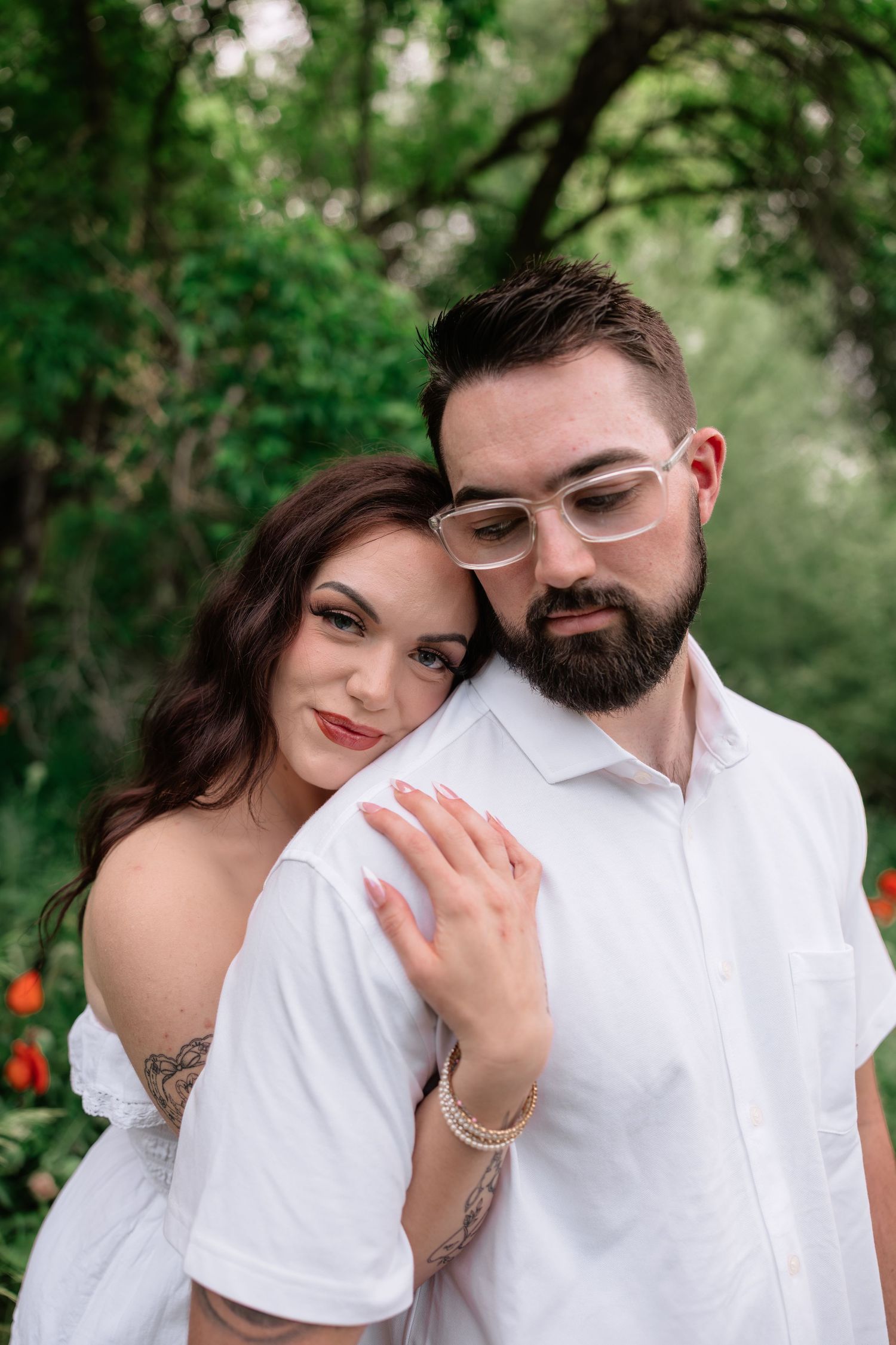 A couple in white outfits embraces outdoors, woman with tattoos leans on bearded man wearing glasses.