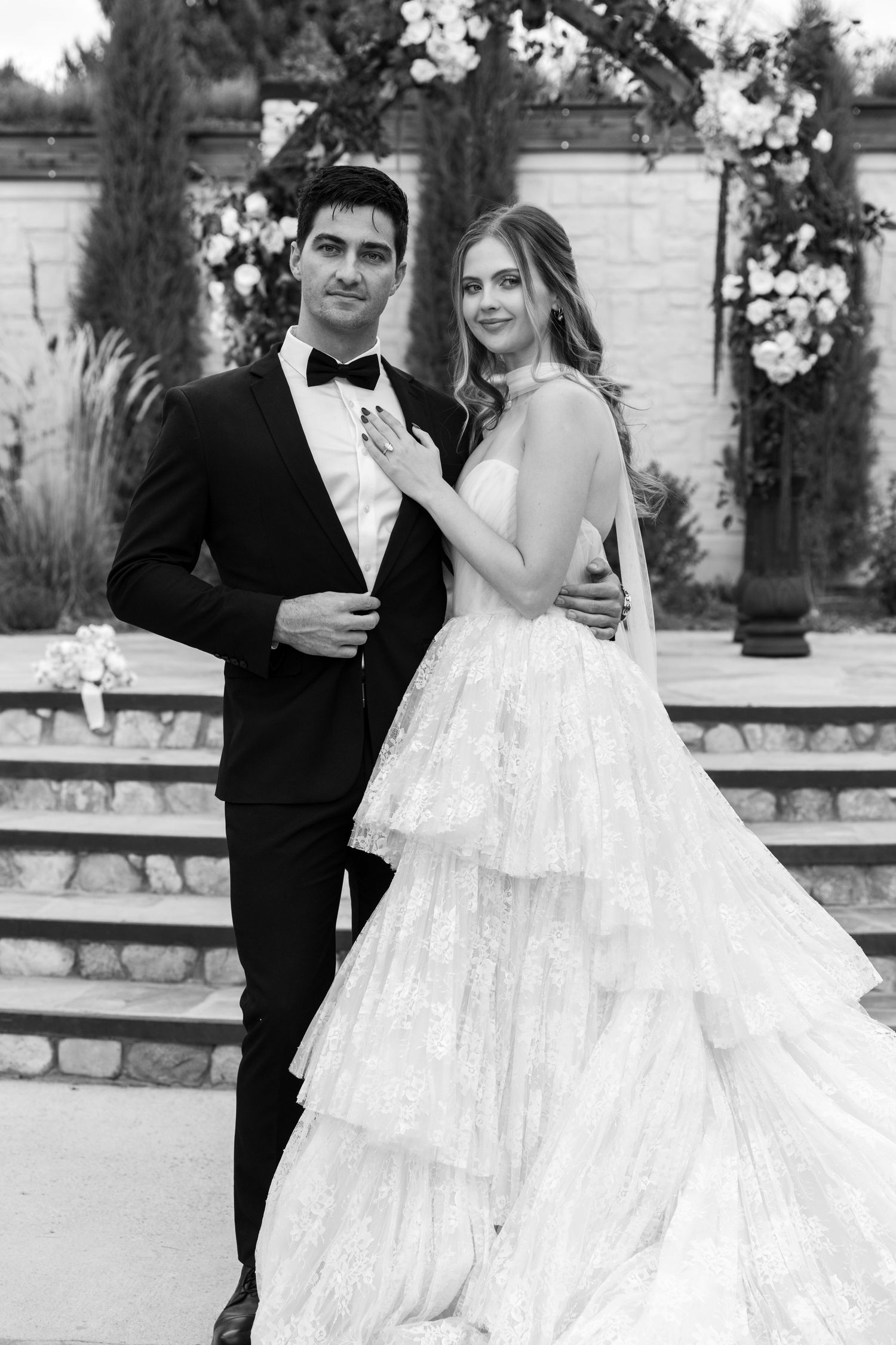Black and white photo of a bride in a tiered lace gown and groom in a tuxedo posing together at an outdoor wedding venue.