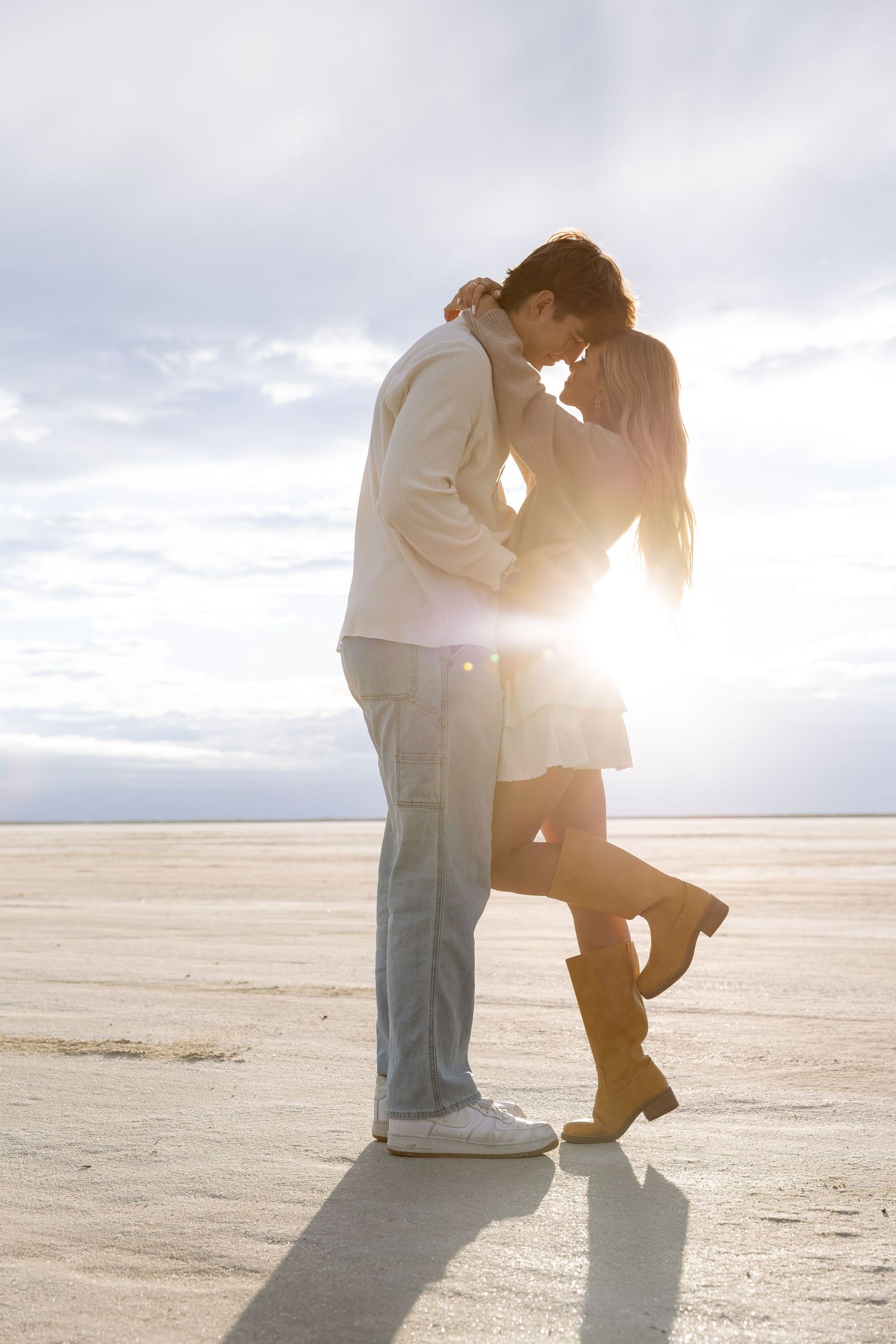 Romantic couple sharing a tender moment kissing on a sunlit beach at sunset with golden backlight.