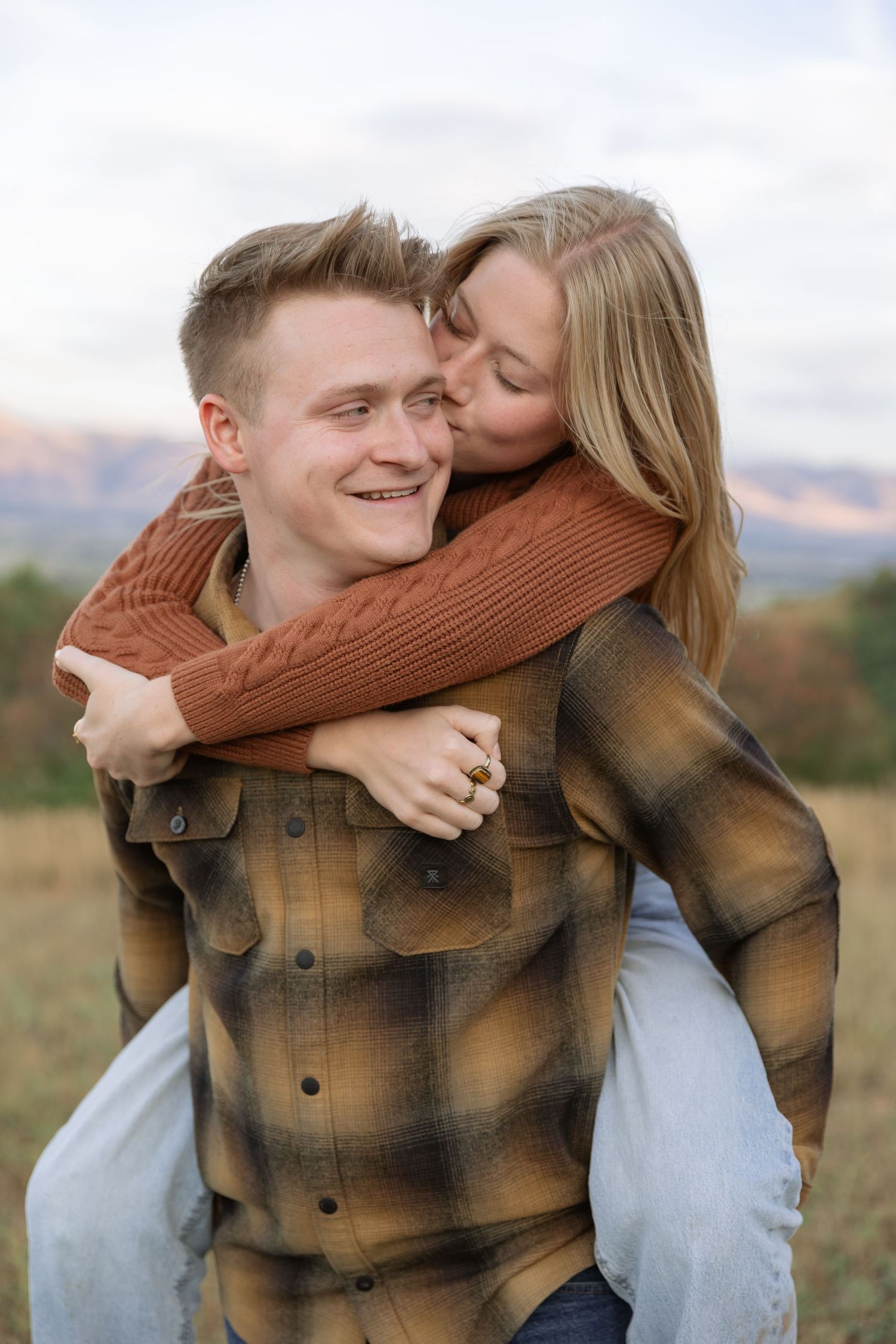 A smiling man gives a blonde woman a piggyback ride as she kisses his cheek in an autumn field.