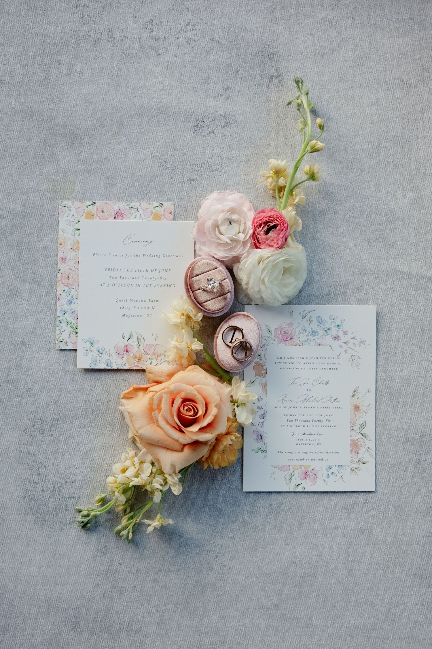 Elegant wedding invitation suite with floral accents, pink roses, ranunculus, and wedding rings on a gray background.