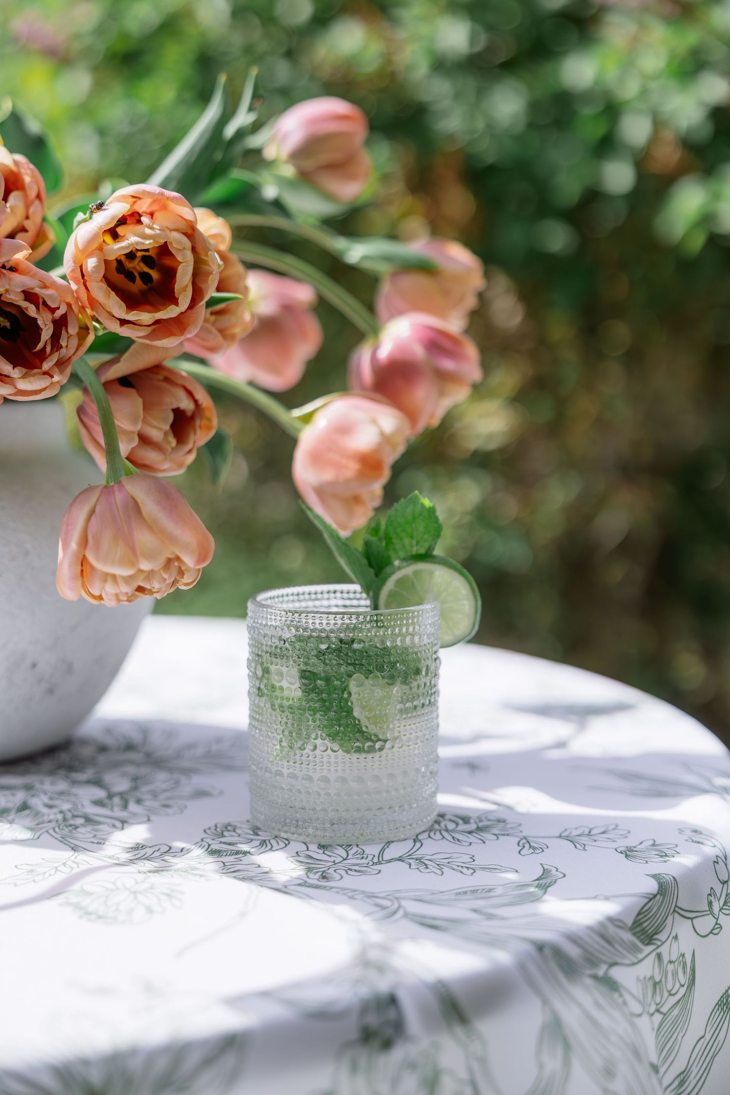 Elegant mojito cocktail with mint and lime beside peach tulips on a floral tablecloth outdoors.