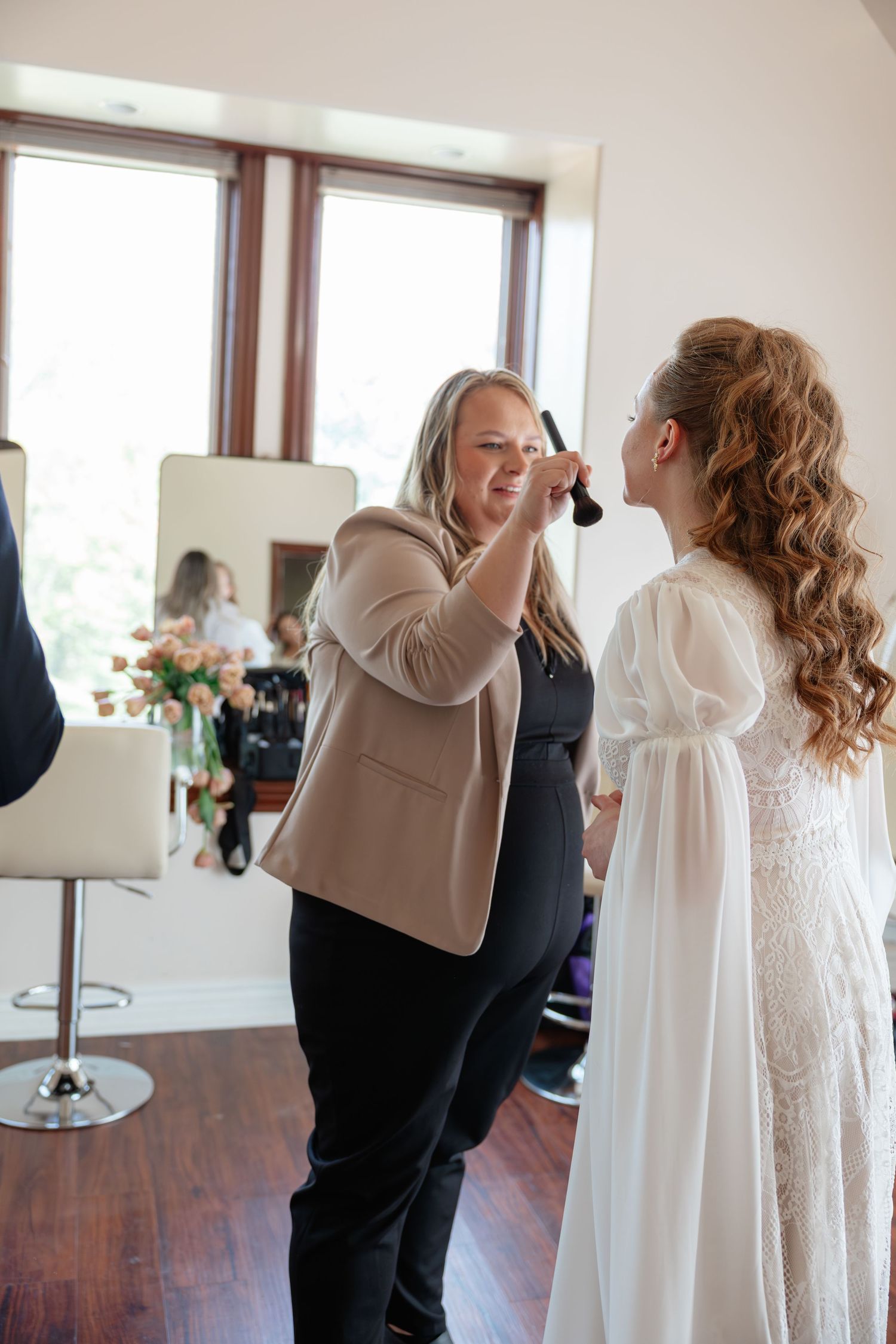 Makeup artist applying finishing touches to a bride in a white lace dress during wedding day preparations indoors.