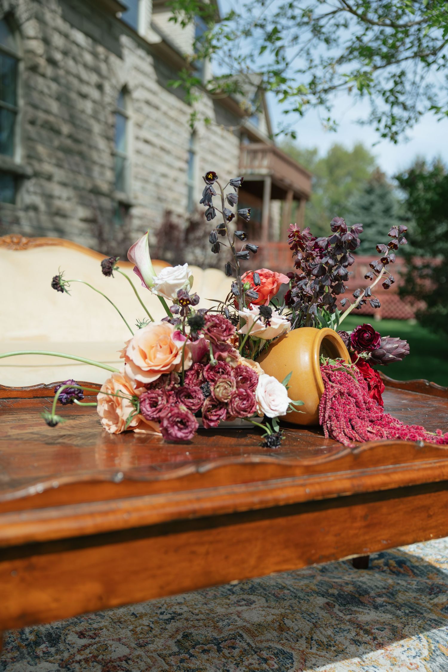Rustic floral arrangement in gold vase with burgundy and peach blooms on antique wooden bench.