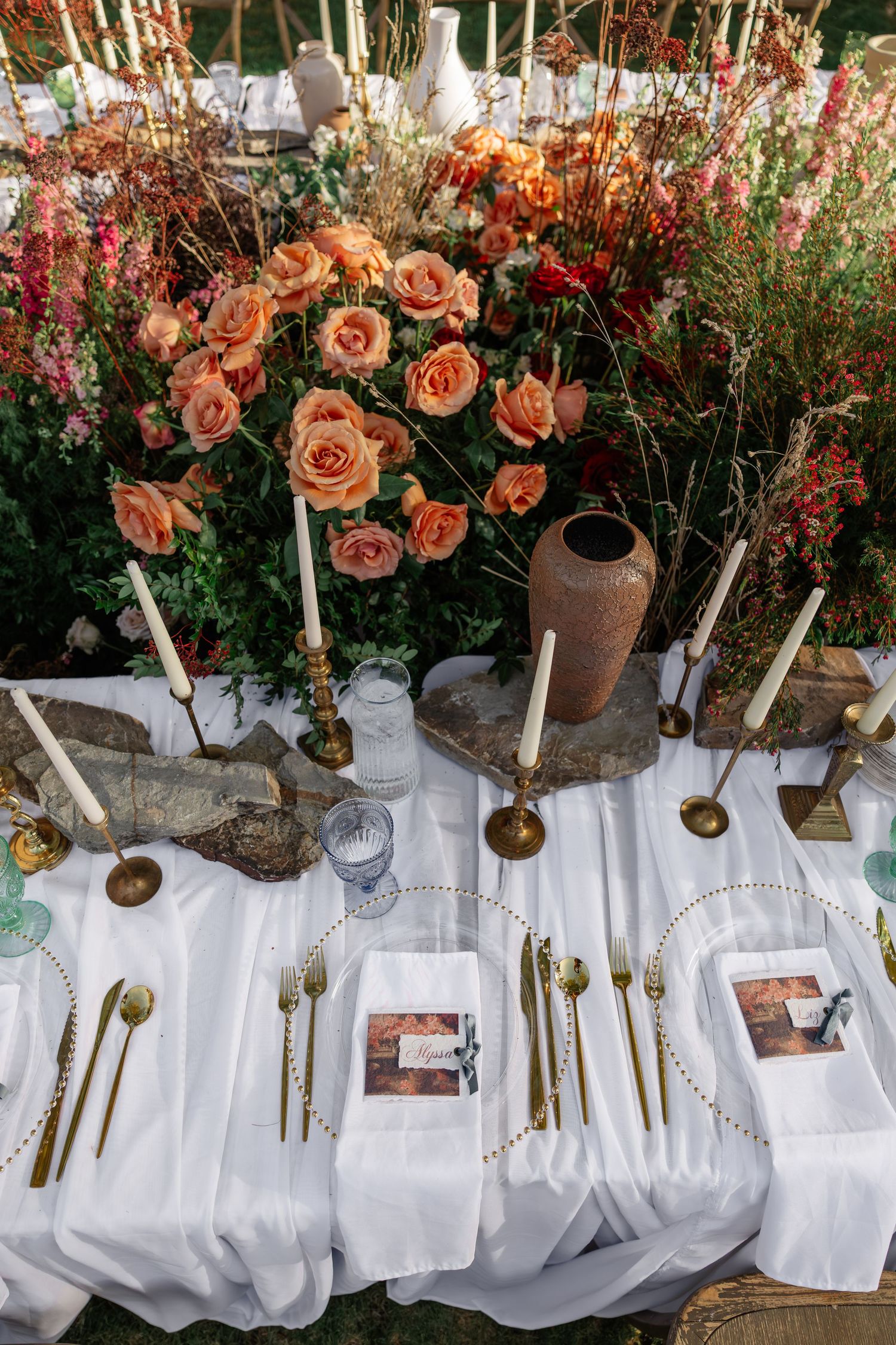 Elegant outdoor tablescape with peach roses, gold cutlery, white linen, and candles for a wedding.