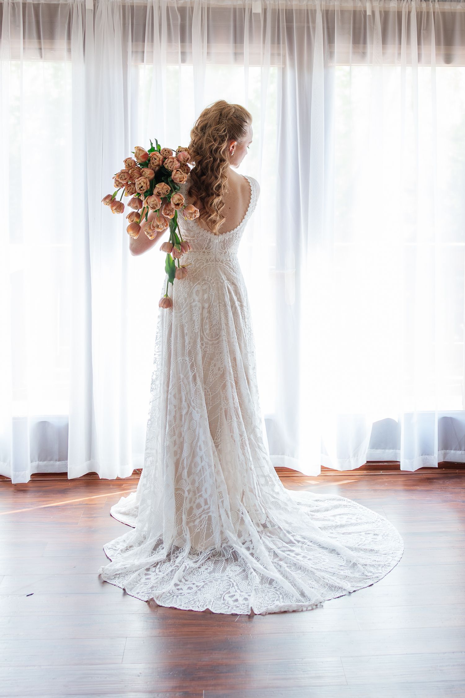 Bride in elegant lace wedding gown with long train standing by window holding peach tulip bouquet from behind.