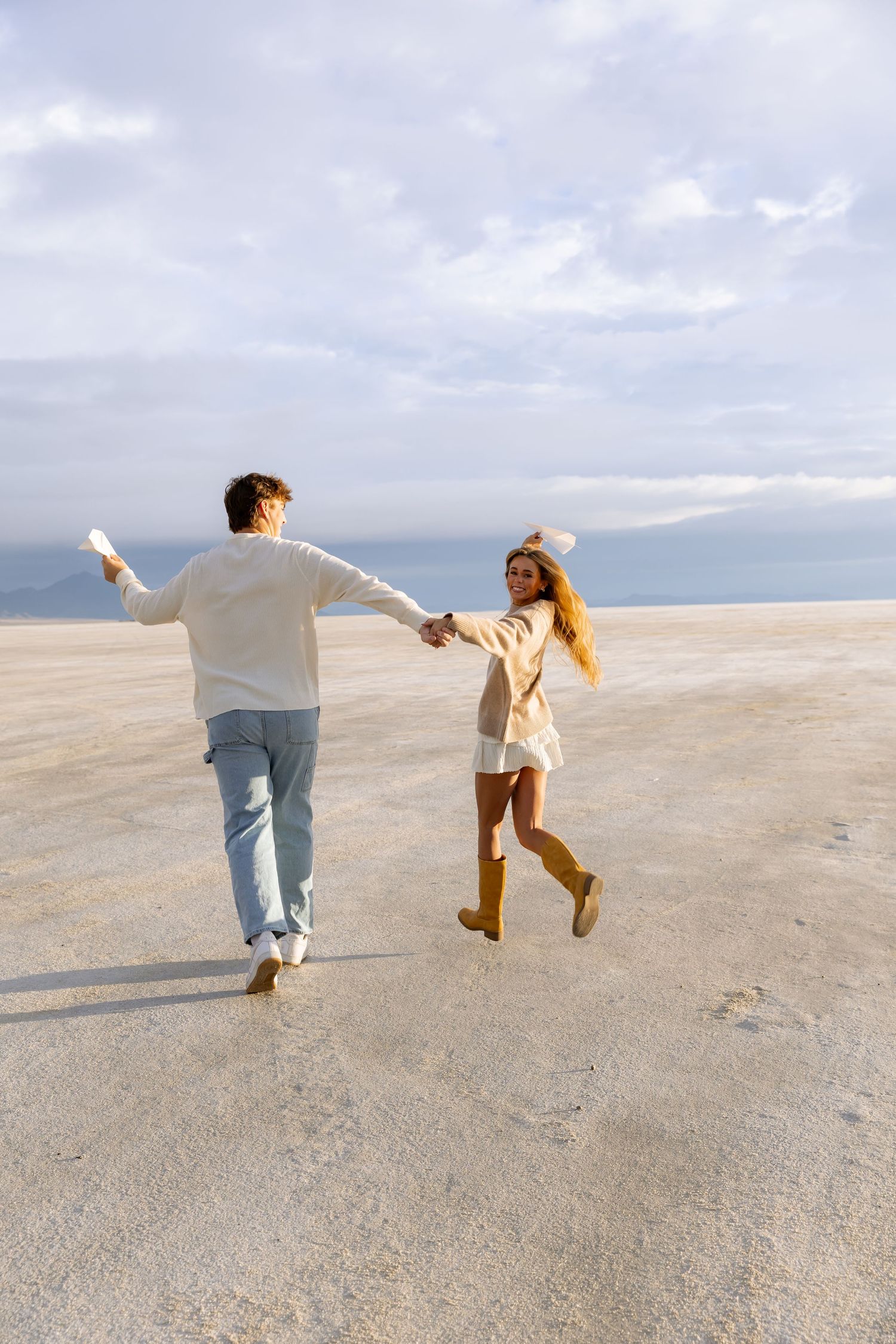 A couple holds hands and runs joyfully across a vast sandy beach under a cloudy sky.