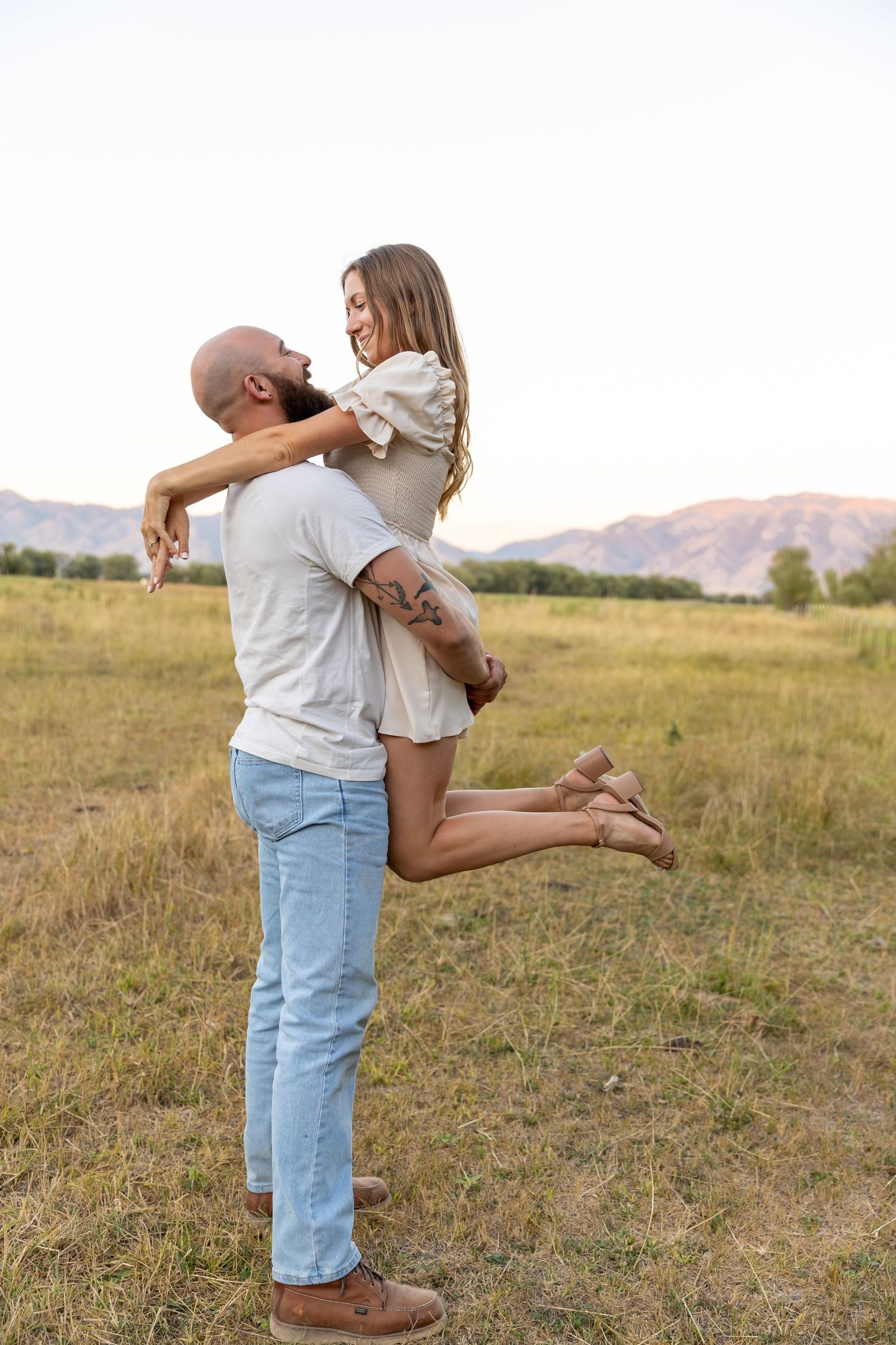 A man lifts a smiling woman in a field with mountains in the background during a romantic outdoor couples photo.