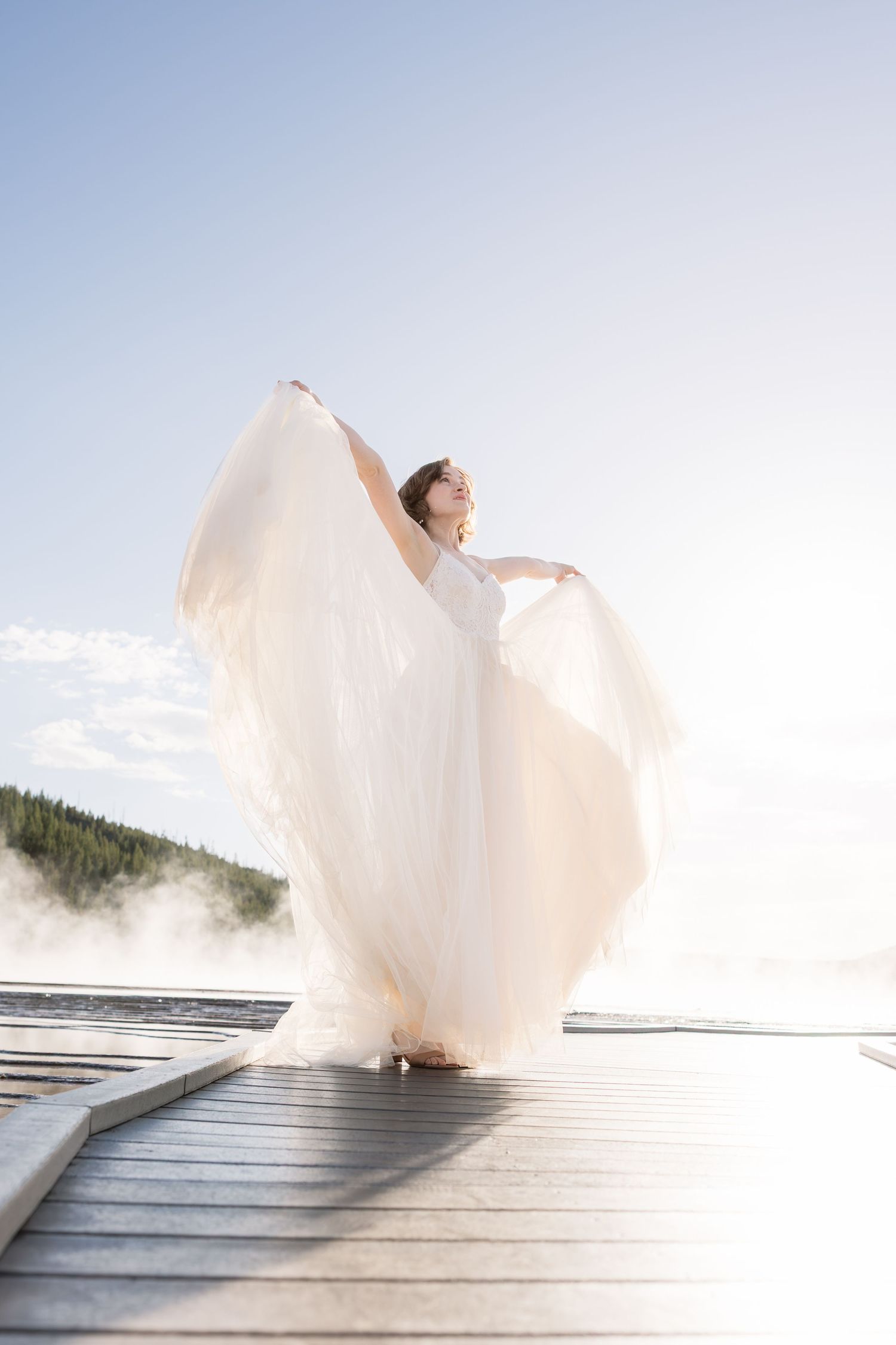 Bride in flowing white gown twirling on a lakeside dock with misty mountains in the background.