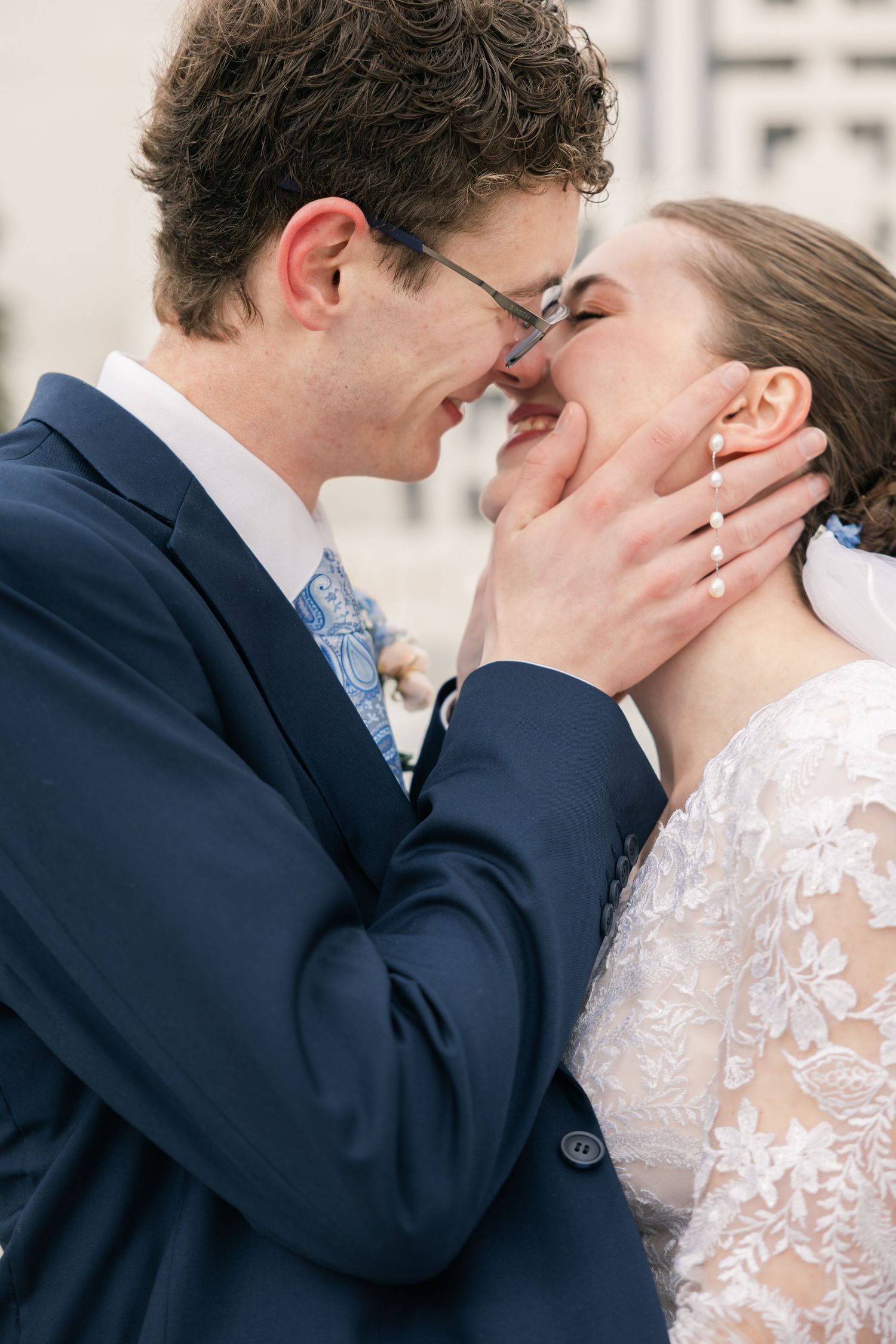A groom in a navy suit tenderly holds his bride's face as they share a romantic kiss on their wedding day.