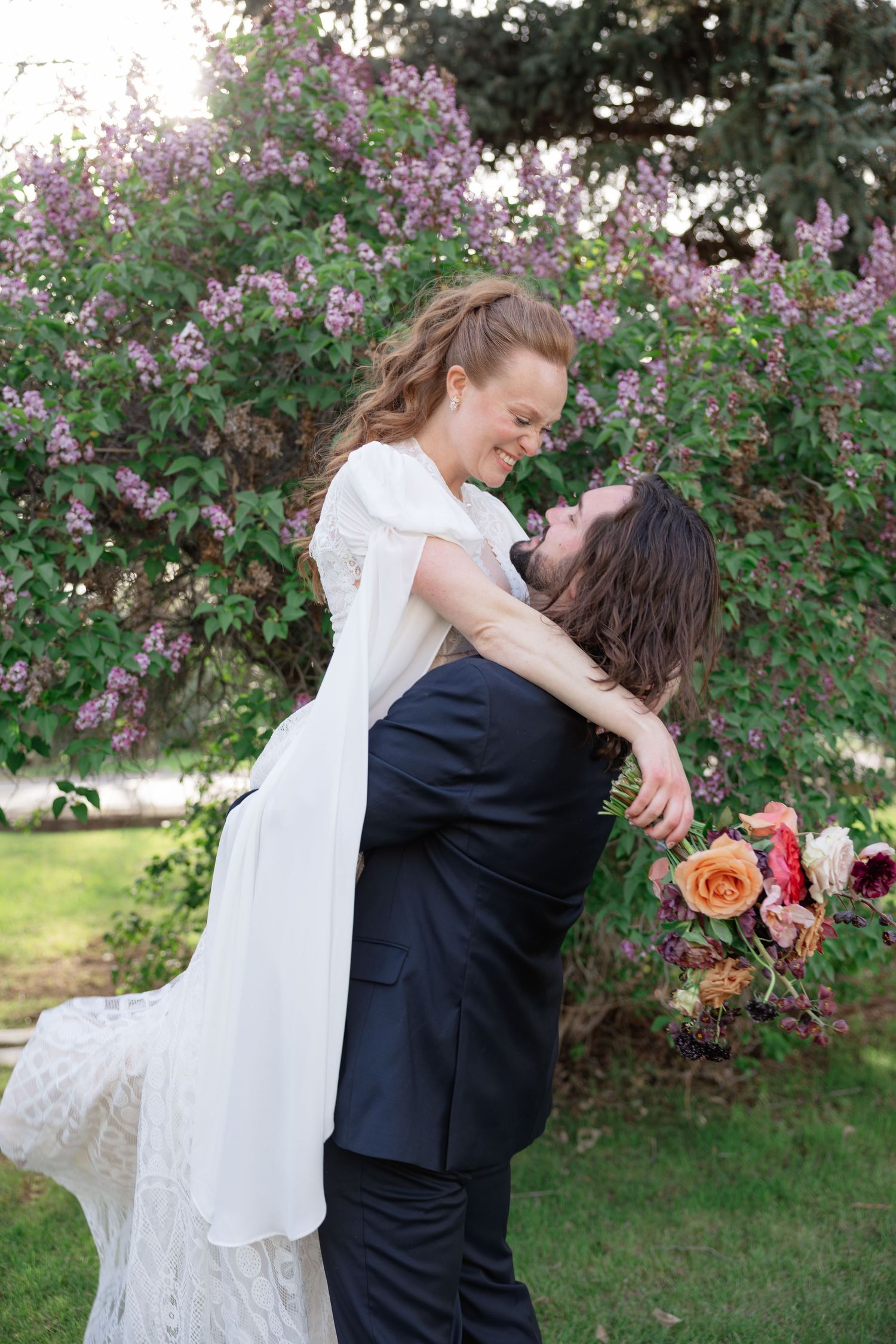 Groom lifting his laughing bride in a lace dress in front of blooming purple lilac bushes outdoors.