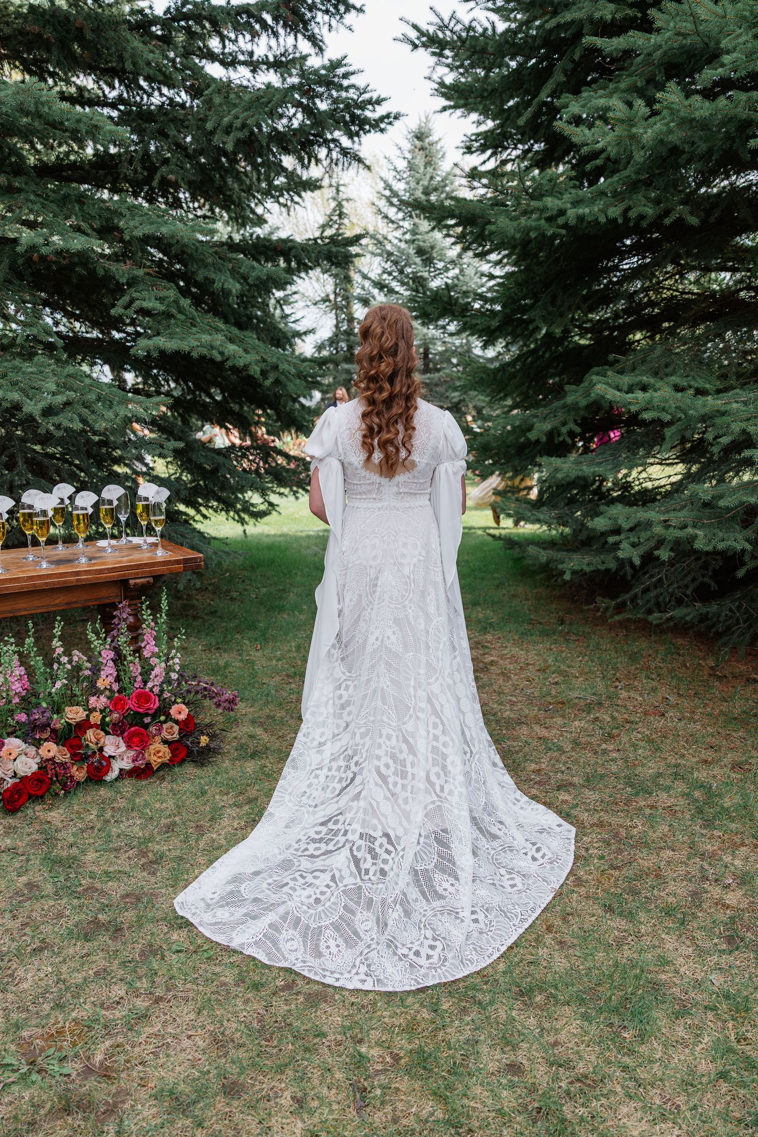 Bride in white lace gown with long train stands facing evergreen trees at outdoor wedding ceremony.