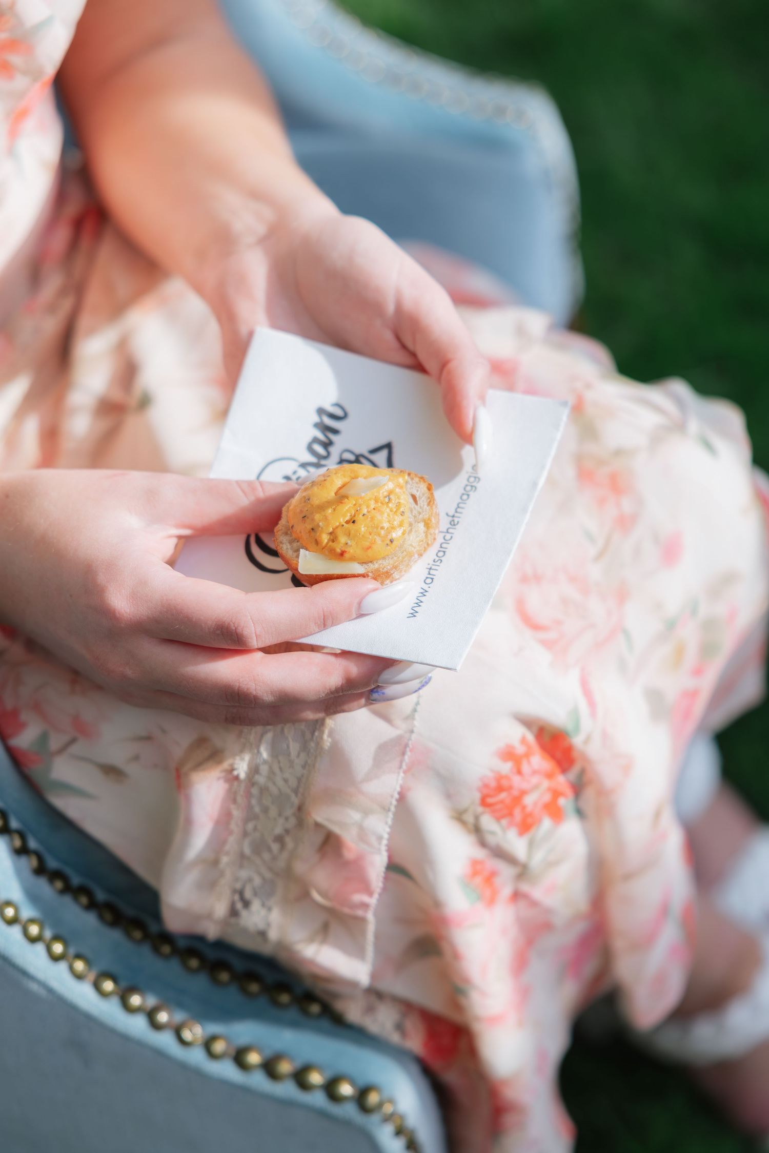 Woman in floral dress holds a small cookie and business card while seated outdoors.