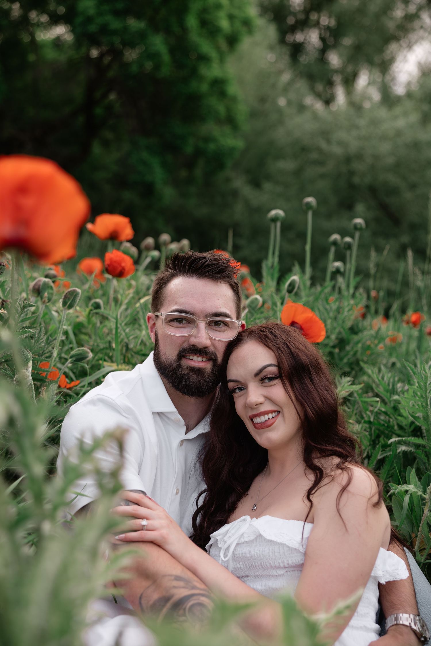 Couple smiling in a poppy field, man with beard and glasses hugging woman in white dress among red flowers.