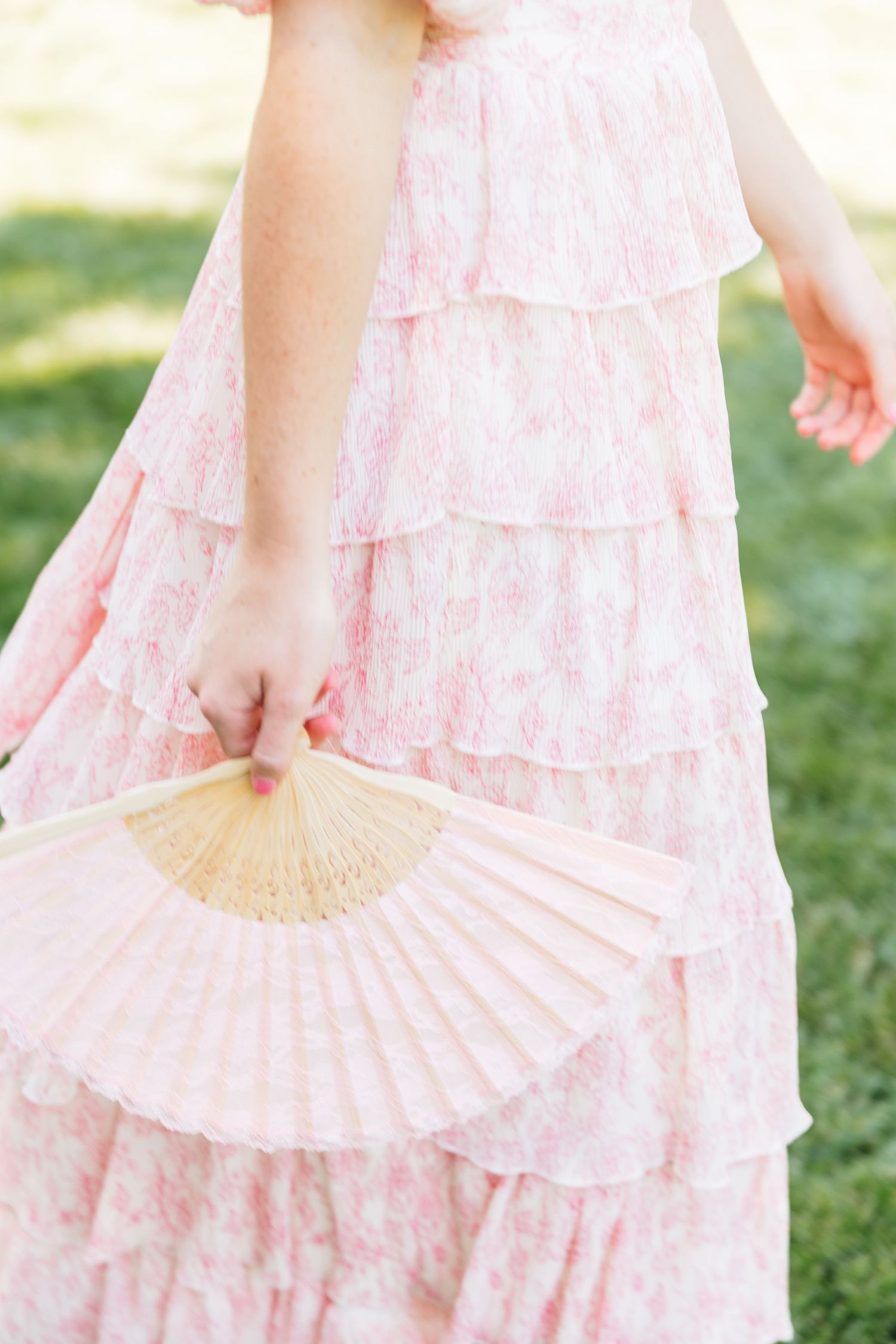 A girl in a pink floral tiered ruffle dress holds a delicate paper hand fan outdoors.