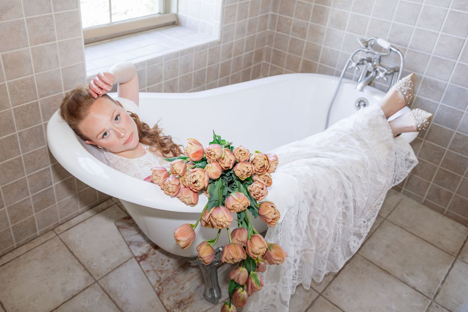 Young red-haired girl in white dress posing in clawfoot bathtub with vase of peach roses in tiled bathroom.