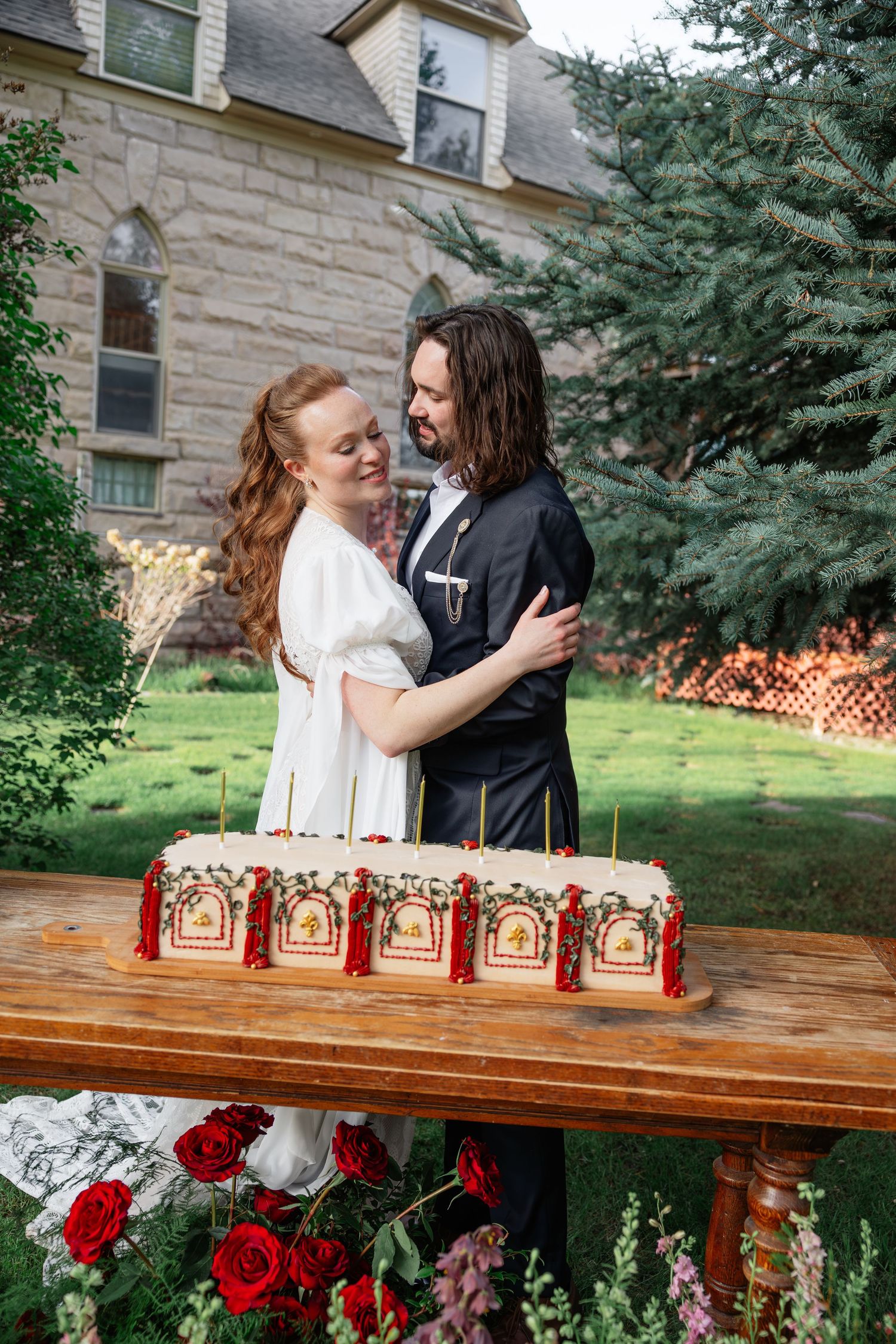Bride and groom embracing behind ornate wedding cake with red tassels on wooden table outdoors.
