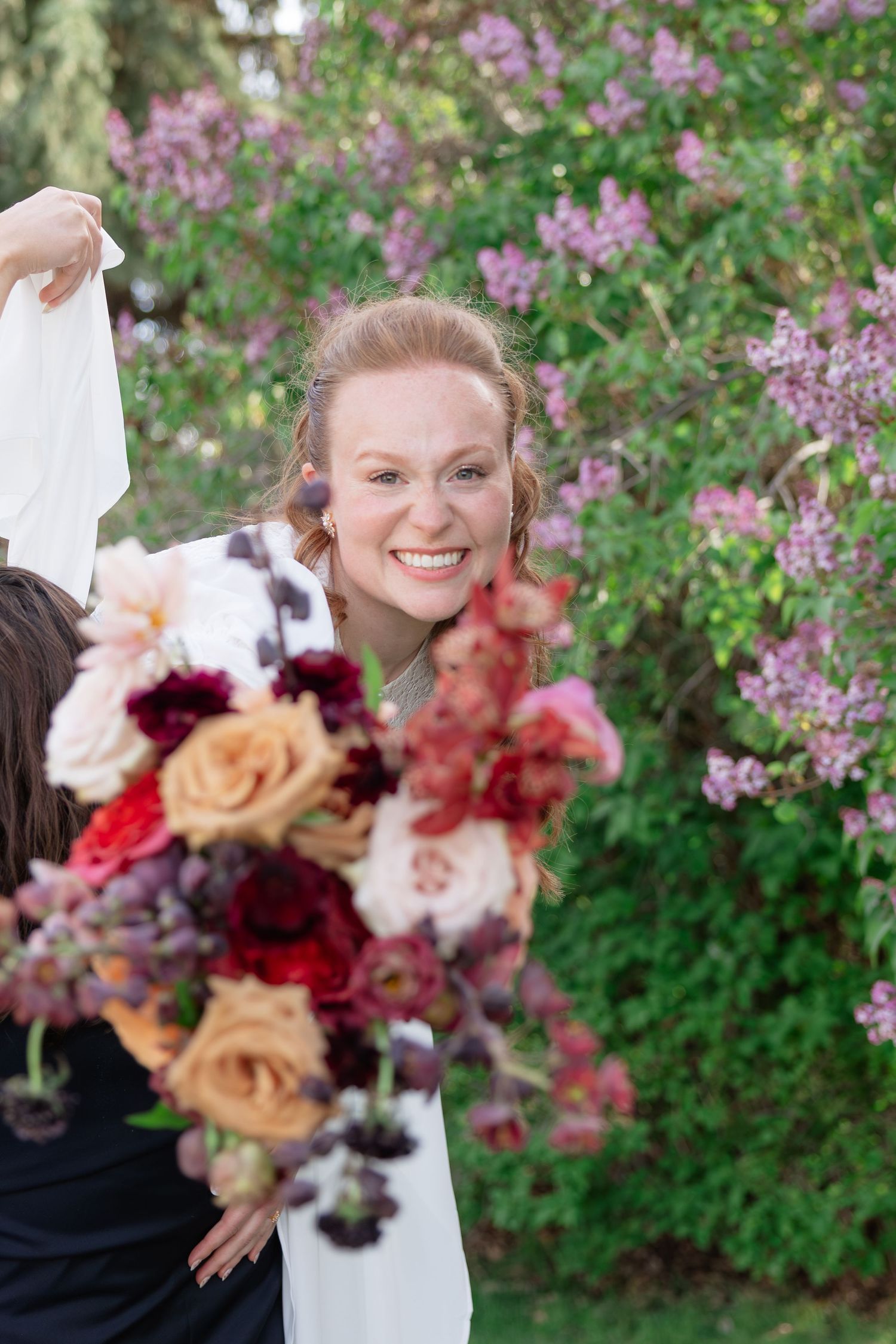 Smiling bride with colorful bouquet of red and peach roses in front of purple lilac bushes.