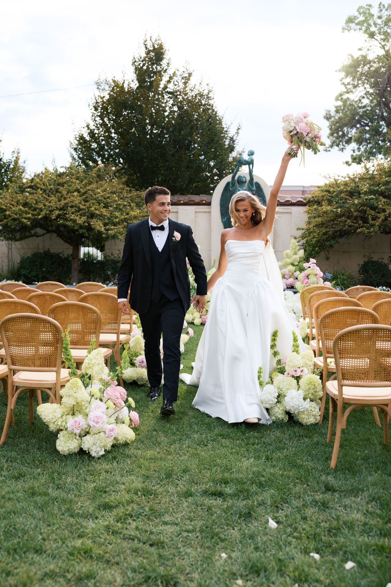 Bride raises bouquet joyfully as couple walks down outdoor aisle lined with pink and white floral arrangements after wedding ceremony.