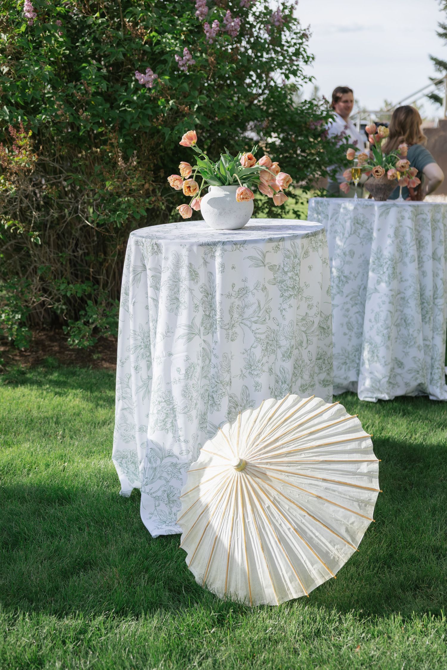 White parasol on grass beside cocktail tables with floral arrangements at an outdoor garden party.