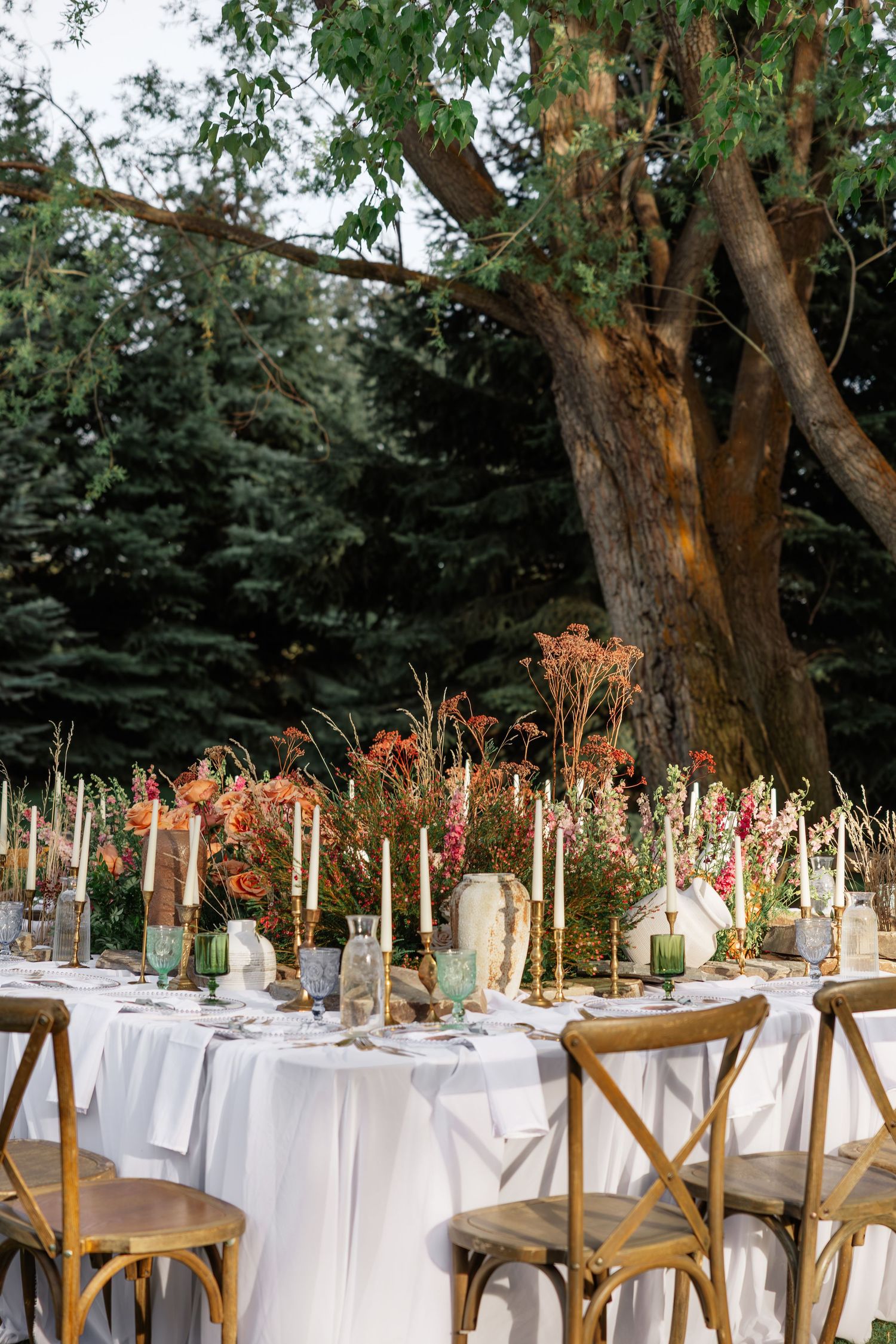 Elegant outdoor wedding reception table with white linens, candles, and autumn floral centerpieces under large trees.