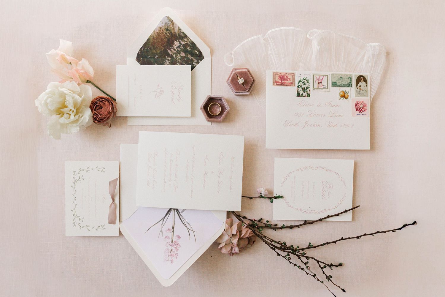 A collection of delicate wedding stationery with floral designs and twigs arranged on a light pink background.