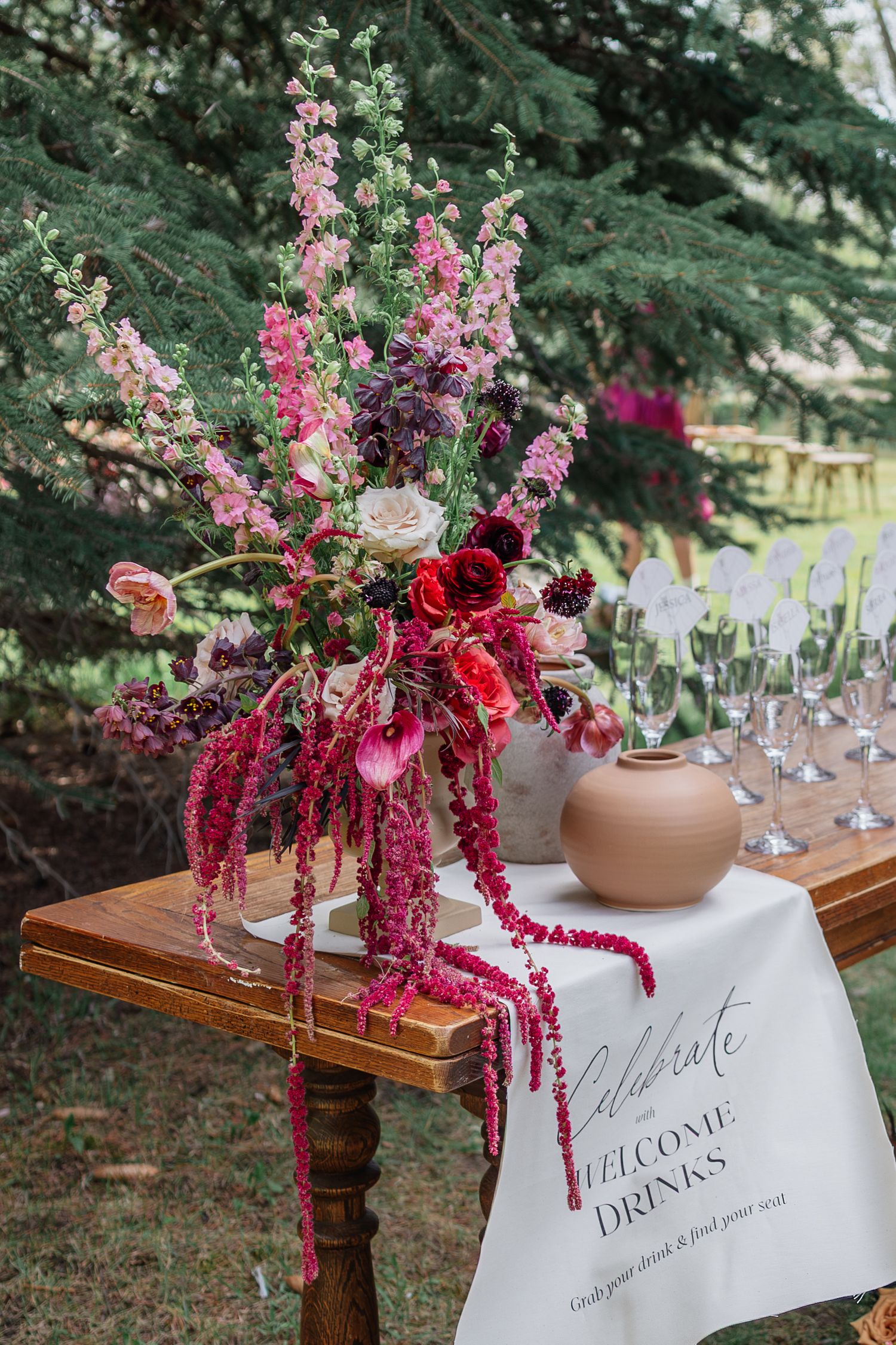 Elegant outdoor wedding welcome drinks table with lush pink and burgundy floral arrangement and champagne glasses.