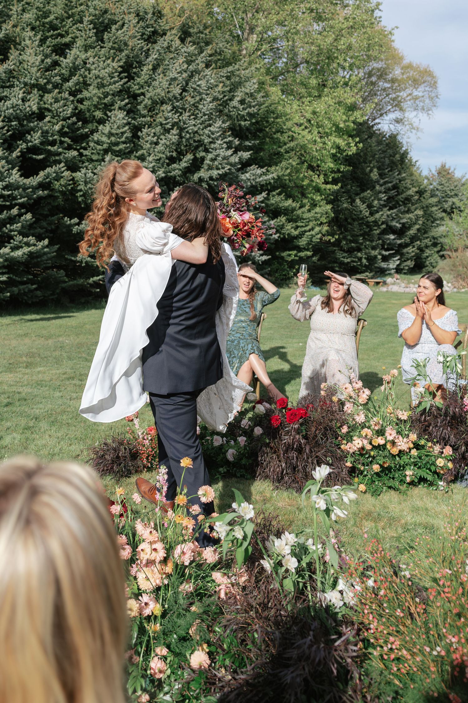 Joyful groom lifts bride after ceremony as guests celebrate among garden flowers outdoors.